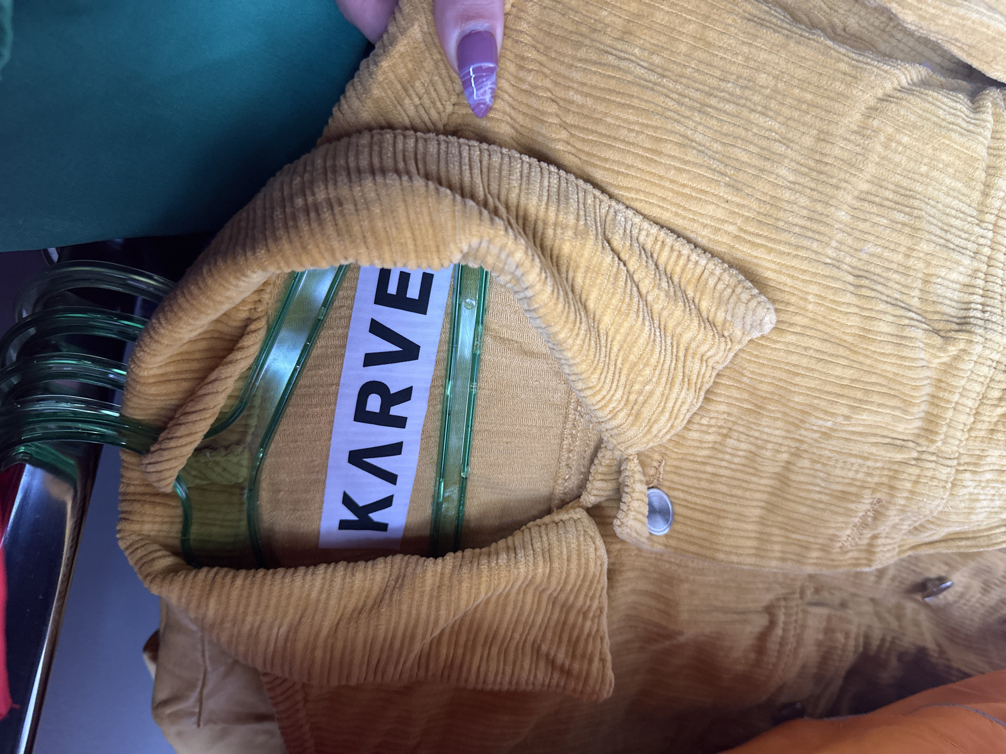 A close-up shows a mustard-yellow corduroy shirt hanging on a green plastic hanger. The hanger displays a white label with the brand name "KARVE" in bold, black capital letters. A hand with a fingernail painted with a lavender marble design is visible holding the shirt's collar. Additional clothing, partially visible, includes a green garment and a metal clothing rack.
There is no indication of this picture's location being the city of Varna, Bulgaria.