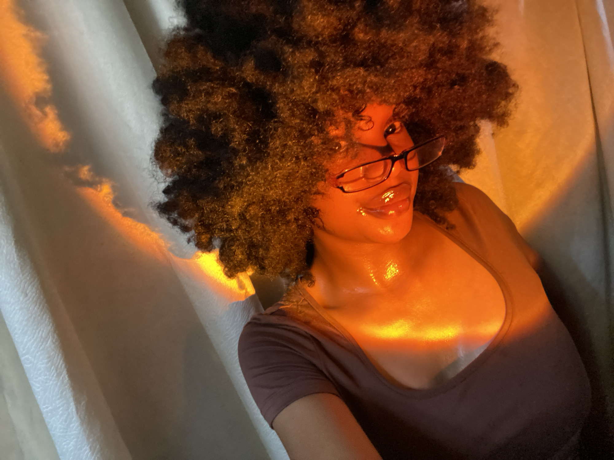 A person with dark skin, wearing a brown t-shirt and glasses, is illuminated by a bright orange light against a white curtain. Their hair is a large, dark brown afro. No information suggests the location is Iranla, Nigeria.