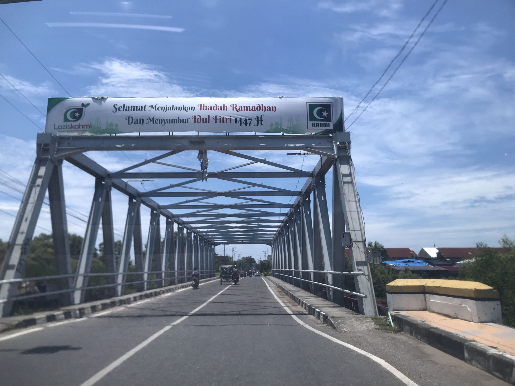 The image depicts a view from a vehicle traveling across a steel truss bridge, likely in Lhokseumawe, Indonesia, based on the banner visible atop the bridge structure. The banner reads "Selamat Menjalankan Ibadah Ramadhan Dan Menyambut Idul Fitri 1447 H," suggesting it is a greeting for Ramadan and Eid al-Fitr. The banner also displays logos for "Lazis Kahmi" and "KAHMI," indicating sponsorship or endorsement by these organizations. The bridge structure is metallic gray, with a complex network of intersecting beams. The road surface is asphalt, marked with white lines. Traffic on the bridge includes motorcycles and a three-wheeled vehicle, possibly a becak. The sky is blue with scattered clouds. On either side of the bridge, vegetation is visible, along with some buildings. The time of day appears to be daytime, with natural lighting.