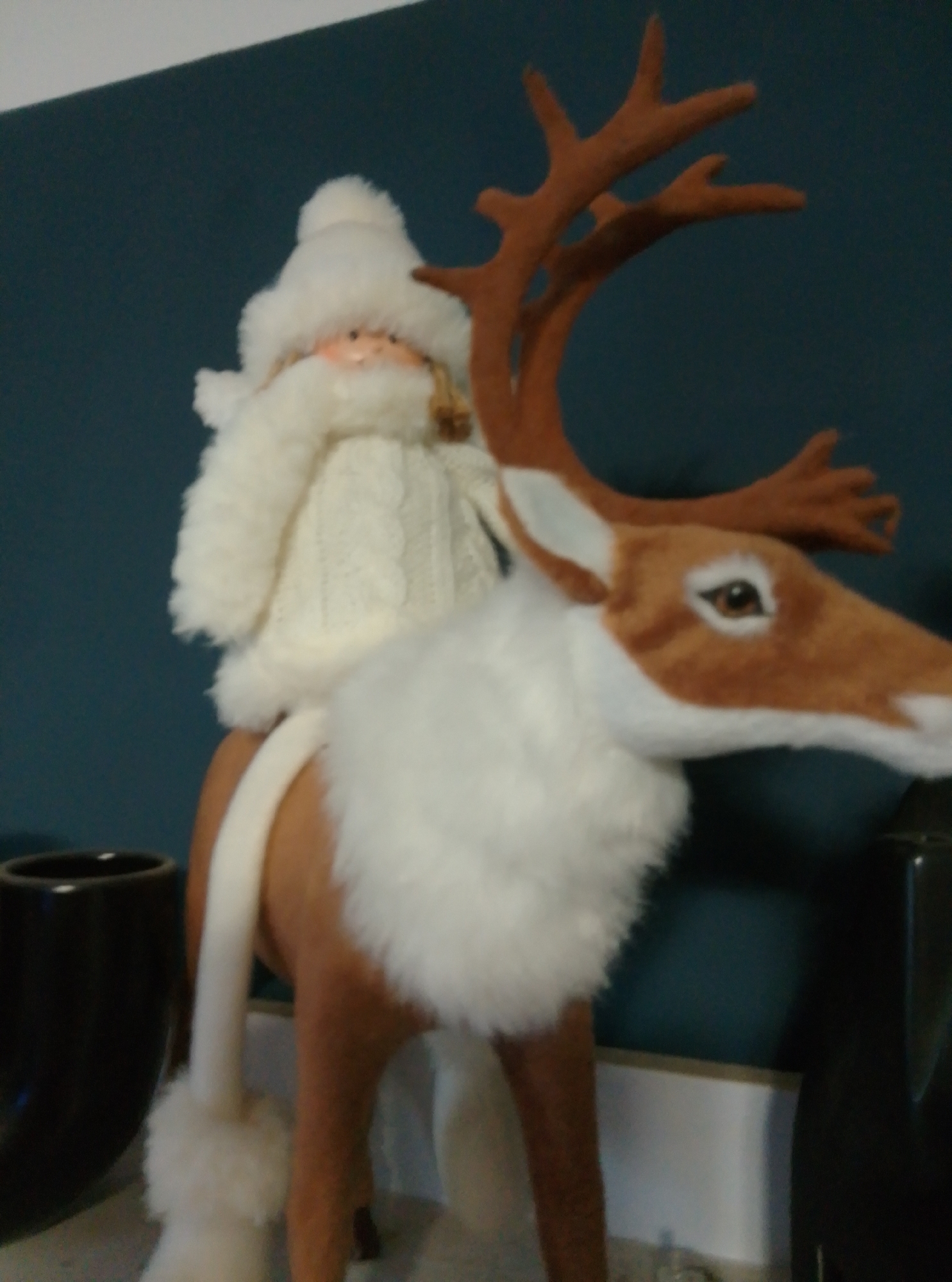 This is a close-up, slightly blurry, eye-level, medium shot of a Christmas-themed decorative item. The item depicts a Santa Claus figurine riding on the back of a stuffed reindeer. The Santa figurine is wearing a white fluffy hat and a white knitted sweater. The reindeer is primarily brown and white, with plush white fur around its neck and a brown head with white fur around its muzzle and ears. The reindeer has brown antlers. The background is a dark teal wall. The foreground shows a portion of a black cylindrical object on the left and a grey surface below. The image does not contain enough information to determine the specific location within Wedel, Germany.