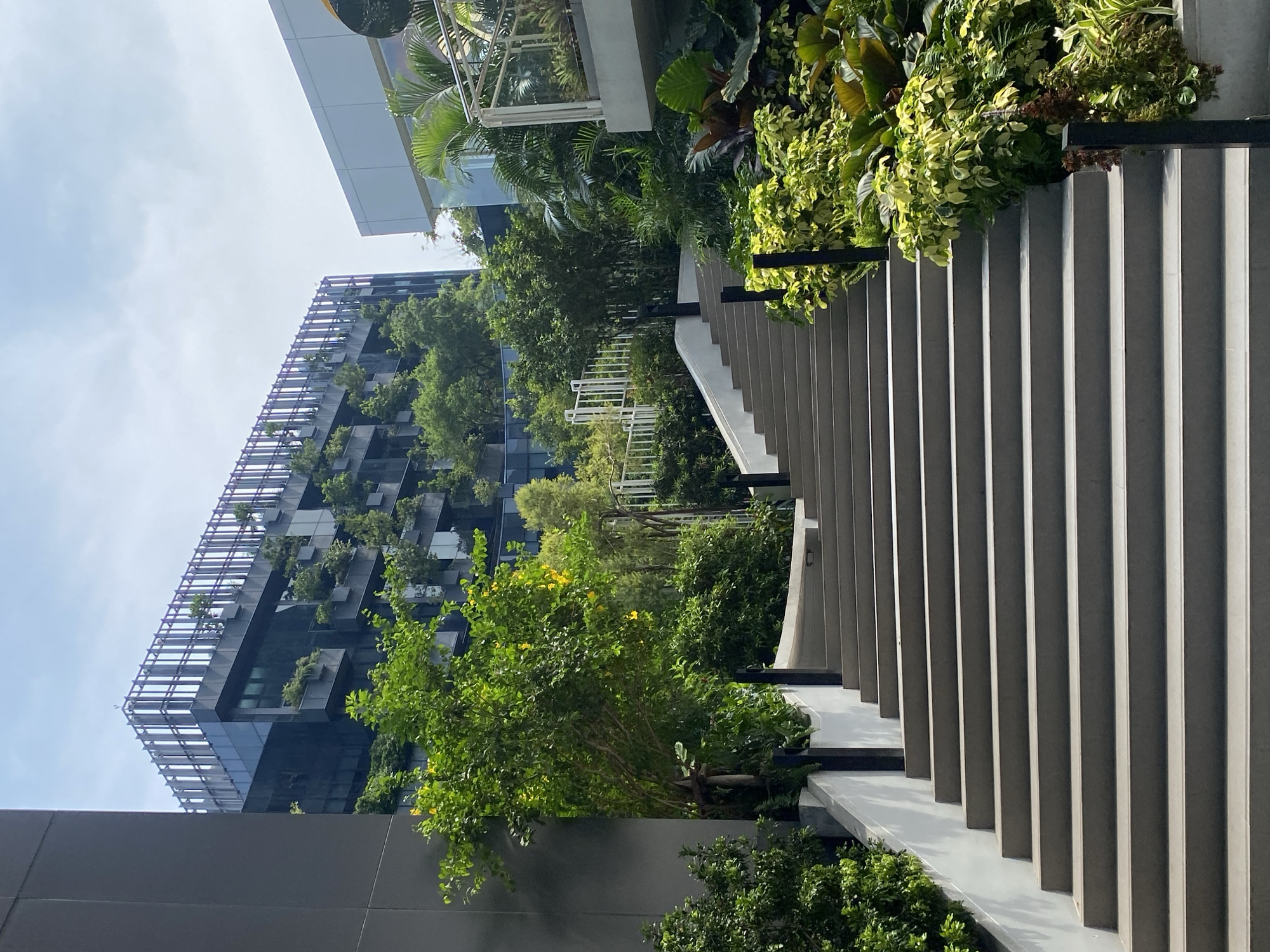 The image depicts a staircase leading up toward a building with extensive greenery, possibly a vertical garden, in Bangkok, Thailand. The staircase is constructed of grey concrete steps with vertical risers, and it is bordered by lush plants and shrubs. The building in the background is a modern structure with multiple levels and extensive green plantings on its facade. The sky is partly cloudy, suggesting a bright day. The photograph is taken from a low angle, emphasizing the height of the building and the ascent of the stairs.