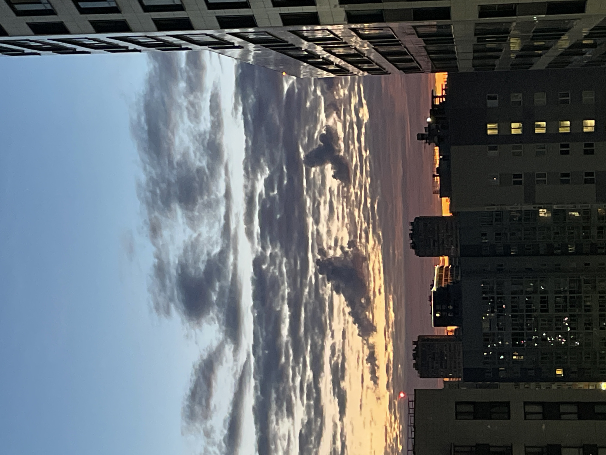 This is an outdoor, eye-level, medium shot of a city skyline at dusk. In the foreground, to the right, is a tall, modern building with many windows, reflecting the sky. To the left, a row of apartment buildings is visible, some with lights on inside. The sky above is a gradient of blues and oranges, with dramatic, dark clouds streaking across. The overall mood is serene and slightly melancholic, with the city lights beginning to twinkle against the fading daylight. The specific location is Unknown, Russia.