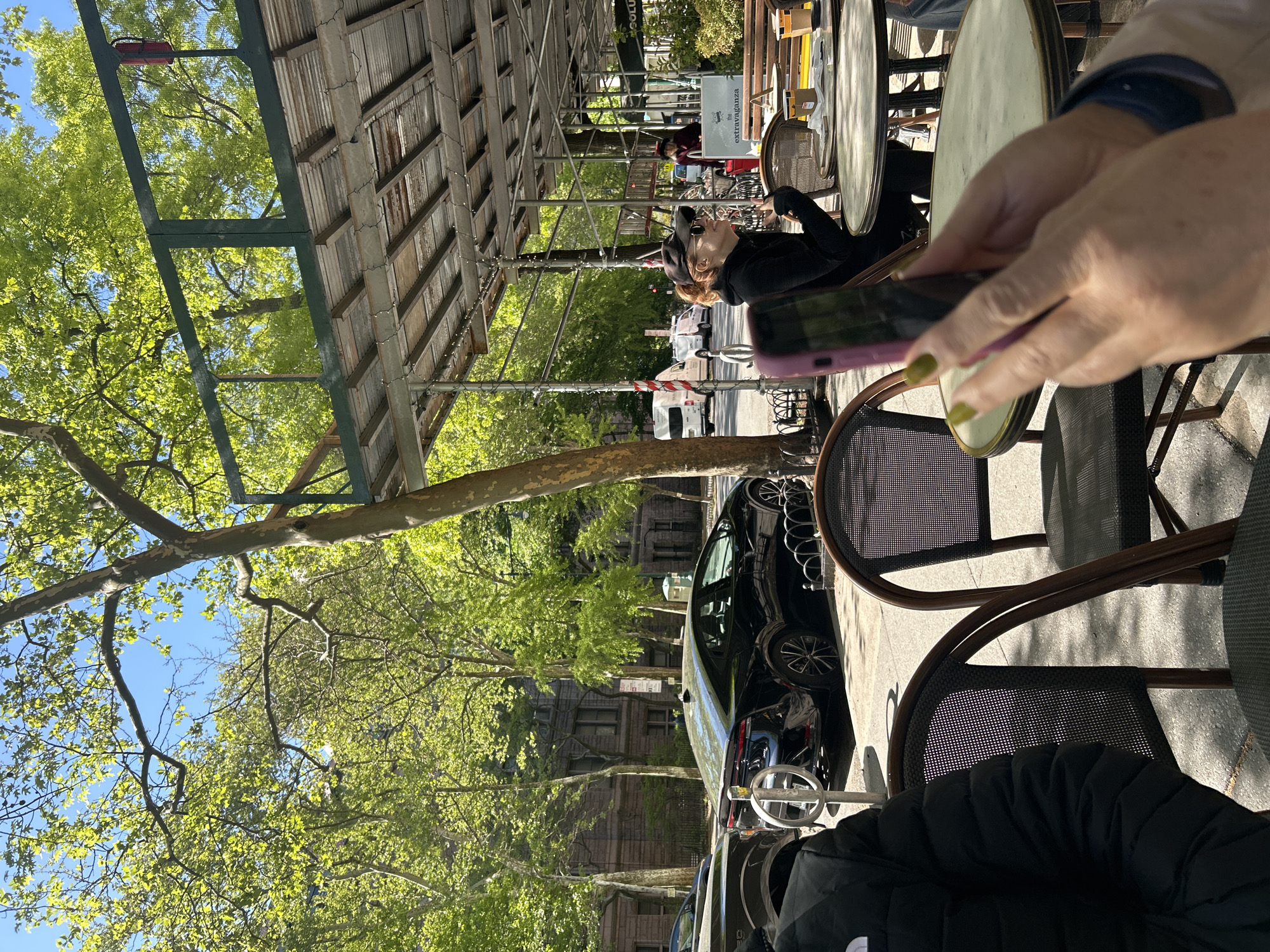 The image depicts a person sitting at an outdoor cafe in New York City, United States of America. The person is holding a smartphone with a pink case. A black car is parked on the street beside the cafe seating area. Other people and cars are visible in the background, along with trees and buildings typical of the city. An awning covers the cafe seating area.