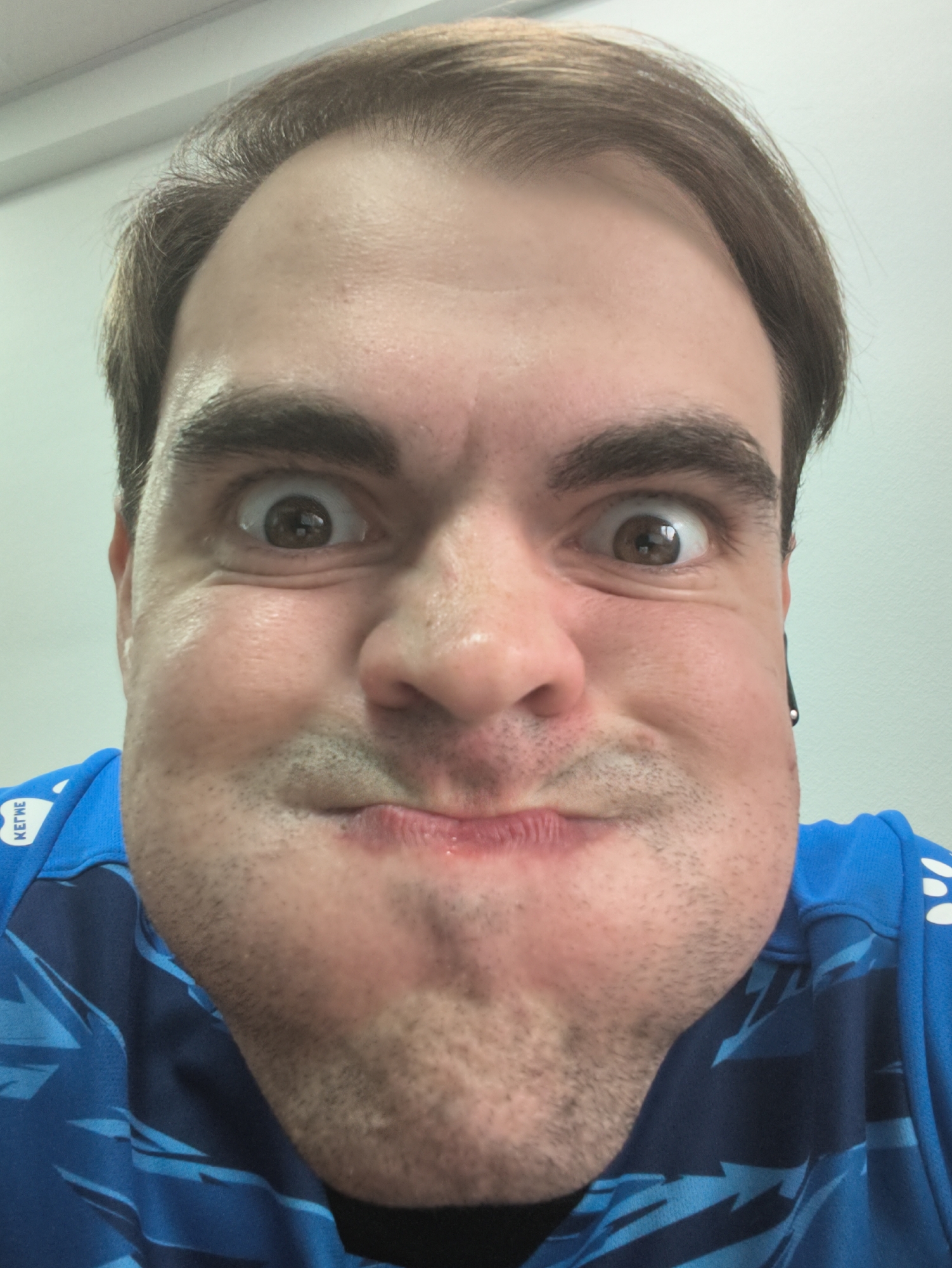 A close-up, eye-level, front-facing selfie shows a man with light brown hair, dark eyebrows, and brown eyes making a puffed-cheek expression. He is wearing a blue shirt with a black and blue pattern and white lettering on the left side of the collar that reads "KETME." The background is a plain, off-white wall. The lighting suggests an indoor setting with the light source coming from the left.