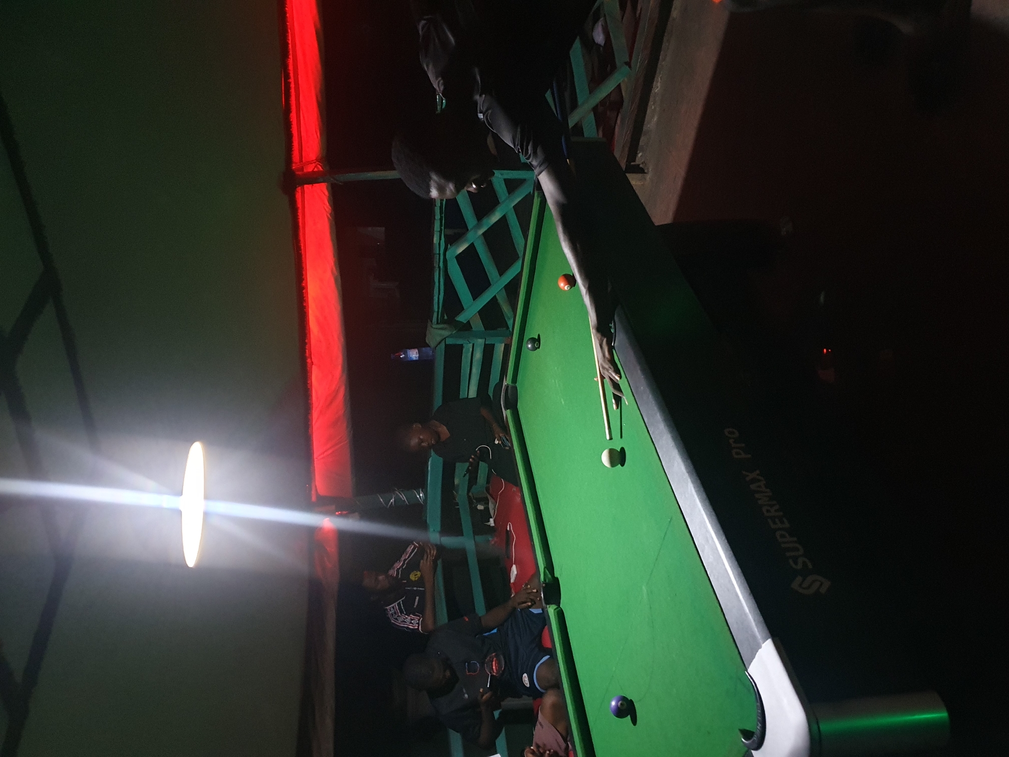 The image depicts a dimly lit outdoor billiards area, likely at a bar or recreational spot, at night. The scene is situated in Lokoja, Nigeria. A bright overhead light illuminates a portion of the green billiards table, where several individuals are present. In the foreground, a man wearing a dark t-shirt is in the process of taking a shot, cue stick in hand, focused on the game. To his left, a purple ball rests on the felt. Further back, another man in a black t-shirt is seated, appearing to be a spectator or waiting his turn. Behind him, another man is visible, wearing a patterned t-shirt and looking towards the table. A bottle of water is visible on a railing in the background, along with red lighting that casts a glow over the upper portion of the scene, suggesting a festive or casual atmosphere. The environment appears to be open-air, with a wooden railing and a corrugated roof structure. The overall impression is of a social gathering centered around a game of billiards.
