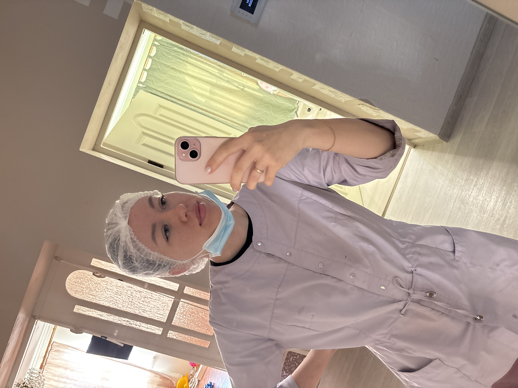 A woman wearing a surgical cap and a blue surgical mask is taking a selfie with a pink smartphone. She is wearing a light purple medical gown. The setting appears to be indoors, possibly in a medical or cosmetic setting. The background includes a doorway and what seems to be another room visible through the doorway, but the location in the city of Kostoryata, Russia cannot be determined from the image.