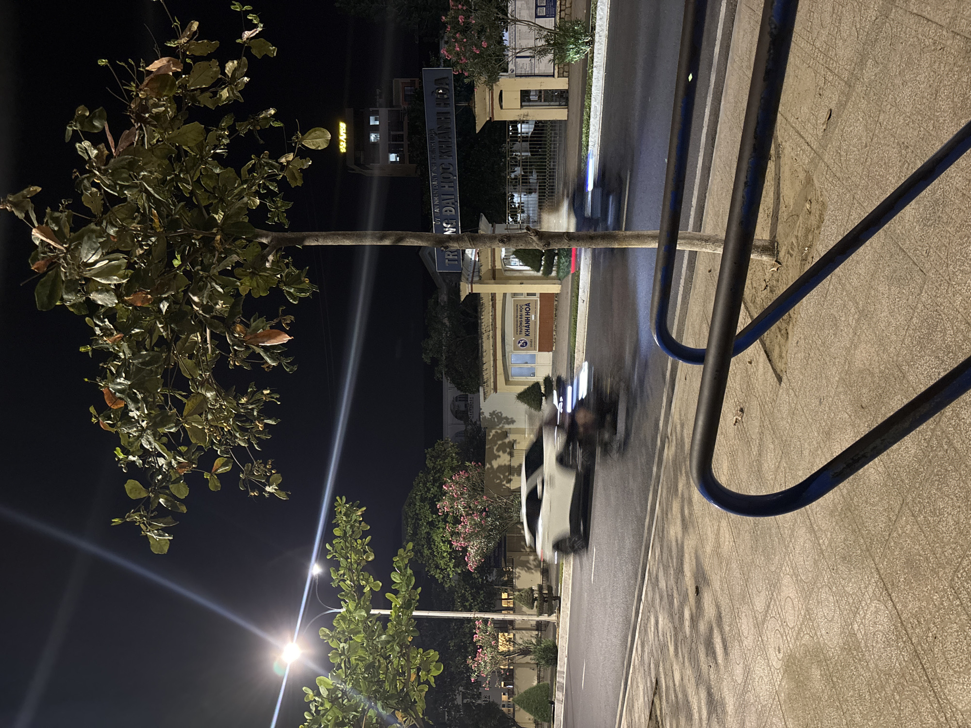 This image depicts a nighttime street scene in an urban environment. A white car is captured in motion on the asphalt road, its headlights illuminating the wet surface. Adjacent to the road is a sidewalk paved with patterned tiles. In the foreground, a dark blue metal railing curves across the lower portion of the frame, casting shadows on the sidewalk.
Behind the car and to the right, a tree with green and brown leaves stands in front of a gate. A large blue sign above the gate reads "TRƯỜNG ĐẠI HỌC KHÁNH HÒA," indicating this is the entrance to Khanh Hoa University. A multi-story building with illuminated windows is visible further in the background, with a sign displaying "922 AVSP" at the top. Another tree with pink flowers is situated to the left of the university entrance. Streetlights are visible in the upper left corner of the image, creating light streaks due to the long exposure. The overall atmosphere is that of a quiet, dimly lit street at night.