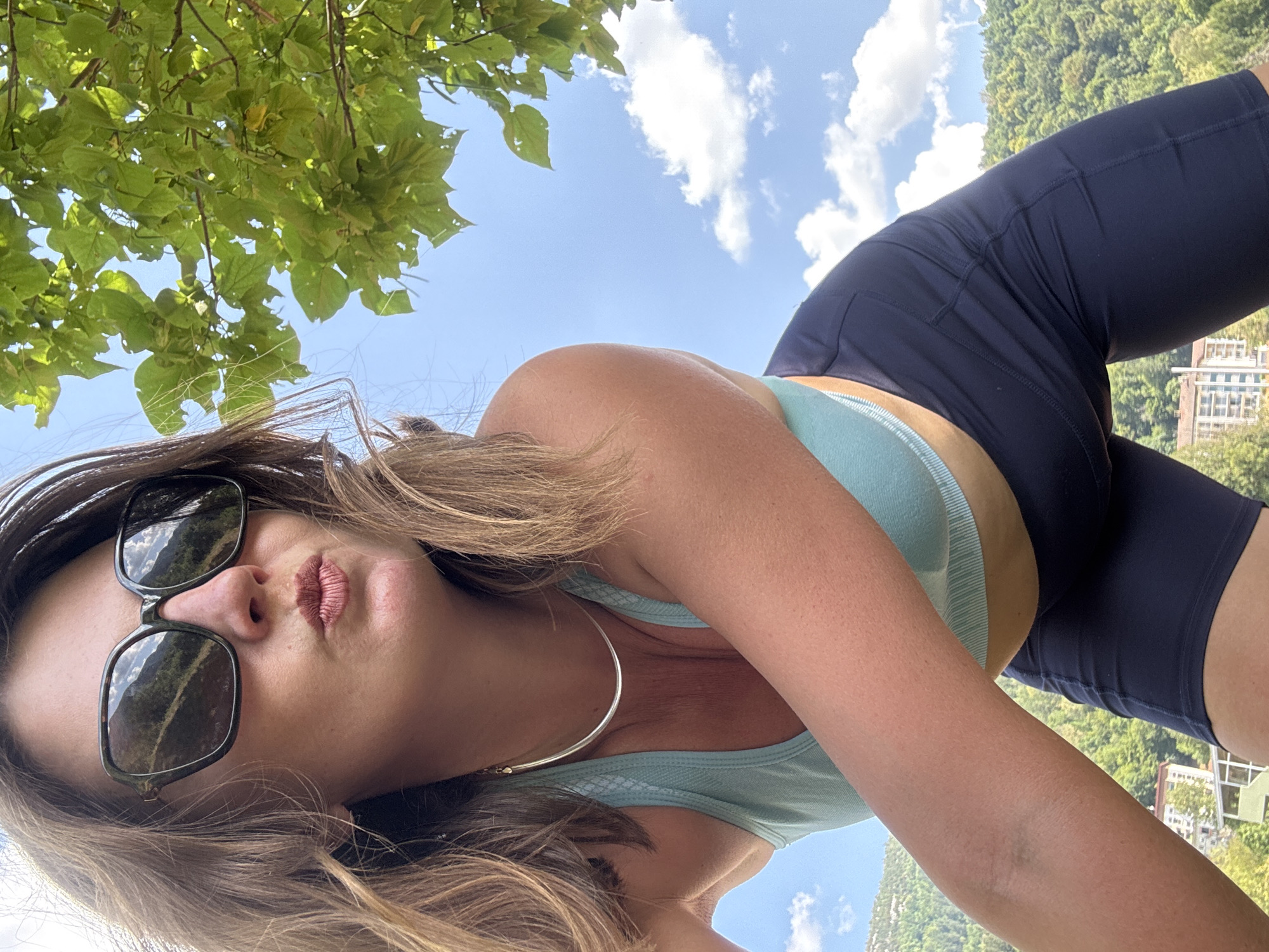 A woman wearing sunglasses and a blue sports bra, leans towards the camera outdoors with a puckered kiss pose. She has brown hair and is wearing a silver necklace. Behind her, a green tree branch with leaves is visible against a blue sky with scattered white clouds. In the background, a lush green hillside with buildings and a body of water can be seen. The scene appears to be in or near Saint-Claude, France.