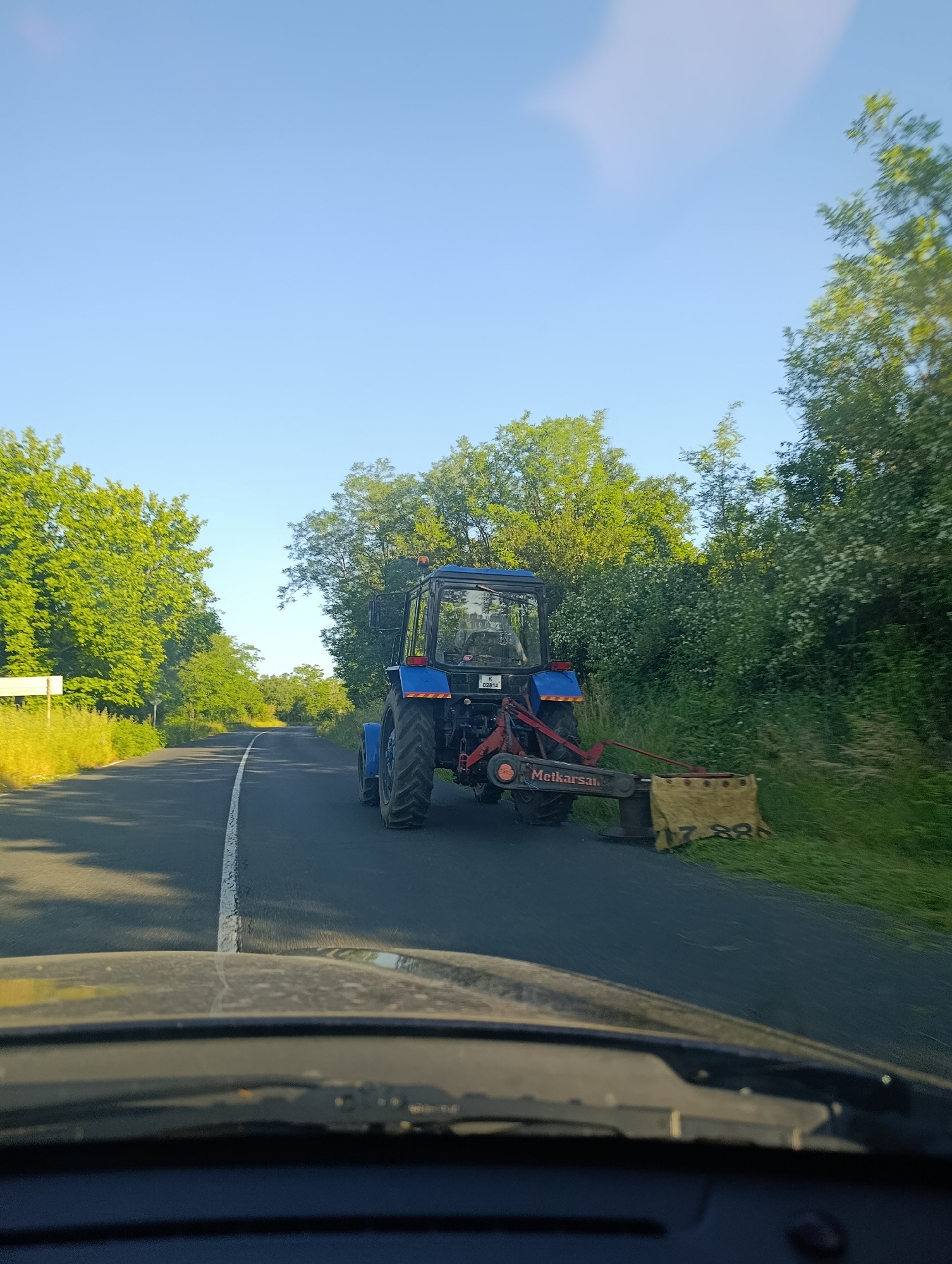 A blue tractor with a rotary mower attachment is driving on a paved road with a white dashed line. The tractor is cutting grass along the side of the road. The scene appears to be in a rural area with trees and vegetation lining the road. The sky is clear and blue, indicating a sunny day. The photo is taken from inside a vehicle, likely from the perspective of the driver, with the dashboard and windshield in the foreground. The license plate on the tractor is partially visible and appears to read "K 02814". The mower attachment has "Metkarsai." written on it. The image depicts agricultural work being carried out in a natural, outdoor setting. The location is identified as the city of Komuniga, Bulgaria.