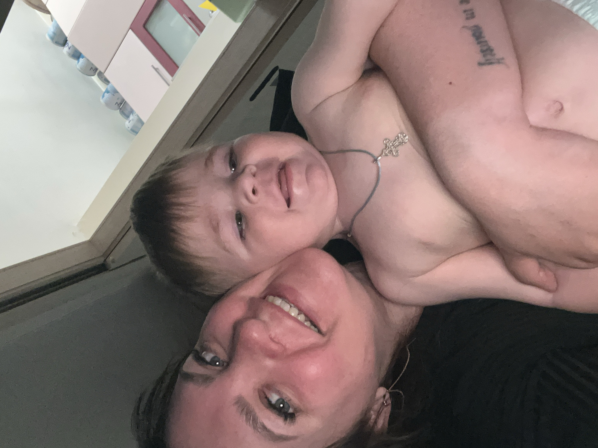 The image shows a woman and a baby taking a selfie. The baby, who is naked except for a necklace with a cross pendant, is being held by the woman. The woman is smiling at the camera. A tattoo is visible on the woman's arm. The background shows a room with furniture. The location is Noviy, Russia.