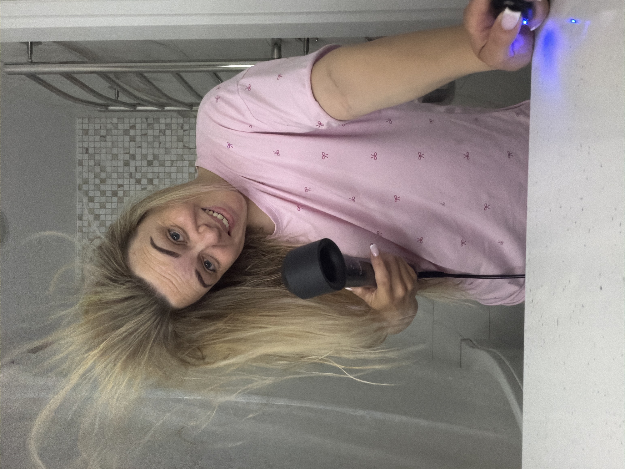 A woman with blonde hair is indoors, likely in a bathroom, holding a hair dryer with a diffuser attachment. She is smiling at the camera. She's wearing a light pink t-shirt with a repeating pattern of small pink bows. In the background, there's a towel rack above mosaic tiles, a white wall, and what appears to be a light switch. One of the woman's hands is holding a device with a blue light on it. The exact location of this scene in Votkinsk, Russia is unknown.