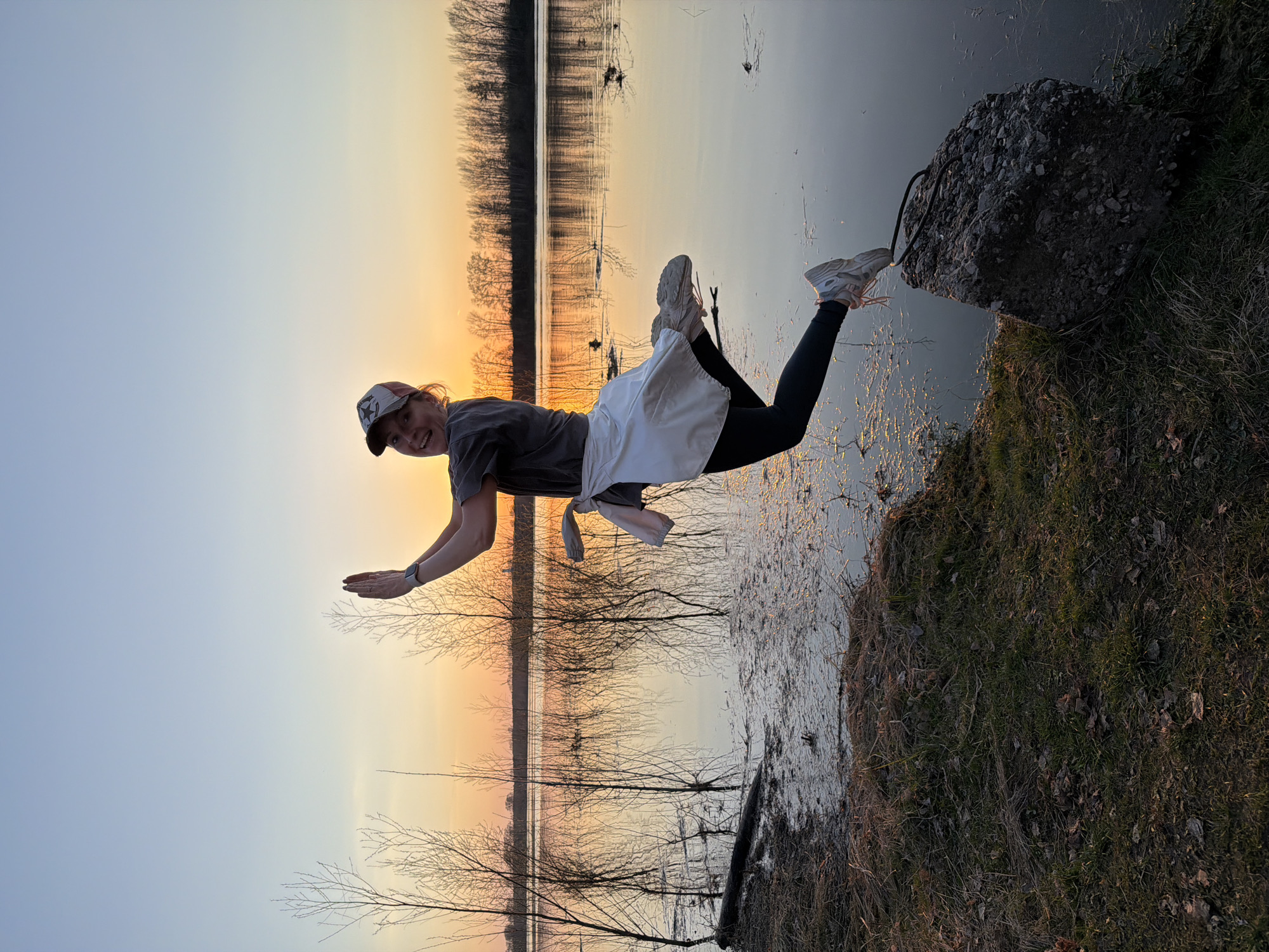 A woman is suspended horizontally in an outdoor setting, seemingly levitating over a body of water at sunset. She is wearing a baseball cap, a grey t-shirt, black leggings, and white sneakers. A white garment is tied around her waist. Her right foot is resting on a large rock. Her arms are extended outwards. The background features a sunset reflecting on the water, with silhouettes of trees. The foreground shows grassy land leading up to the water's edge. The image was taken in the city of Chaikovsky, Russia.