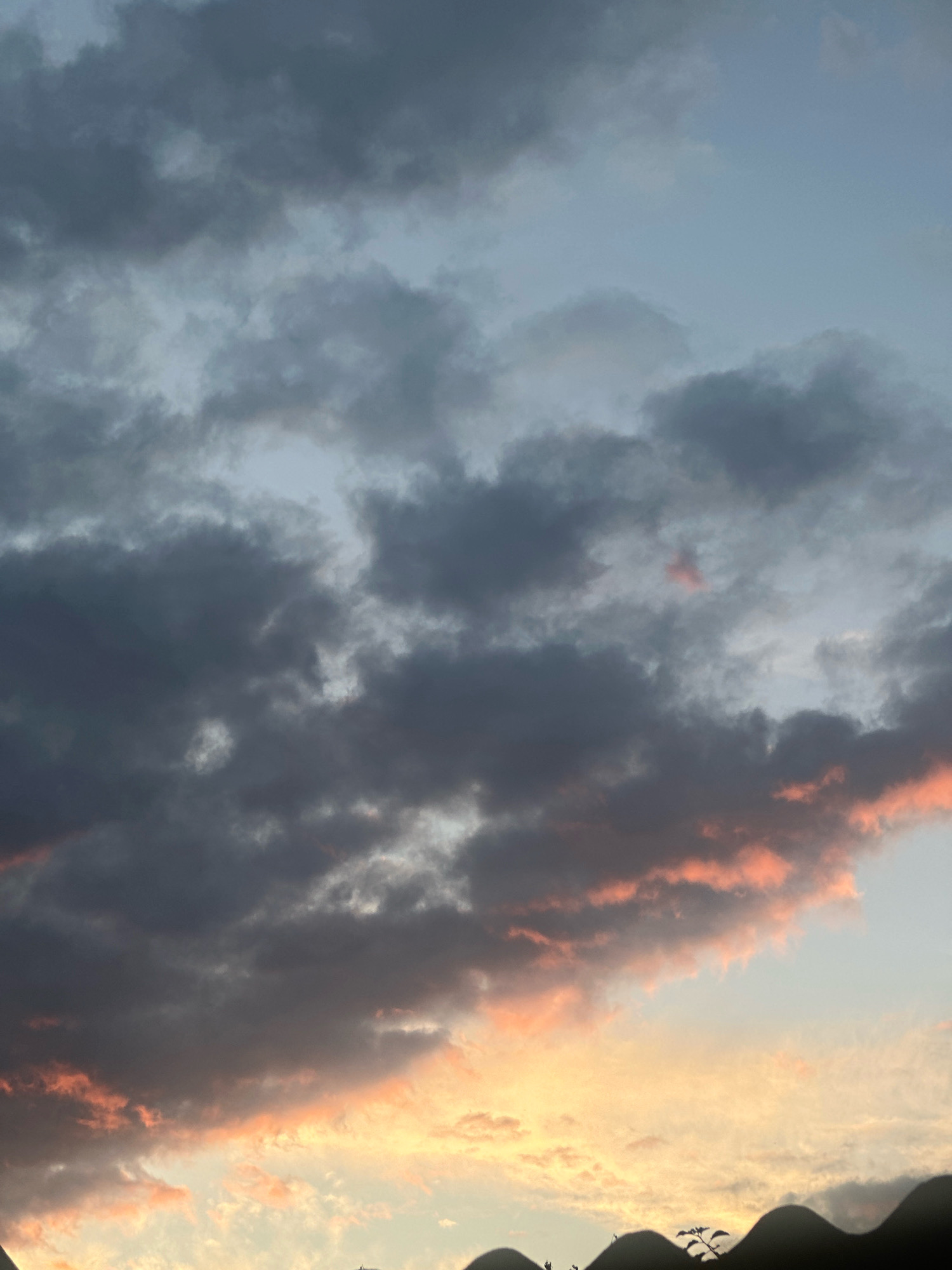 A low-angle shot captures a cloudy sky at sunset over Хацюхоўскі сельскі Савет, Belarus. The clouds are a mix of dark grays and lighter blues, with streaks of pink and orange visible where the setting sun is breaking through. The bottom of the frame is dominated by the dark silhouettes of undulating rooftops, likely belonging to residential buildings, with a small plant or tree silhouetted against the sky on the right. The overall mood is dramatic and slightly somber due to the heavy cloud cover and the dimming light.