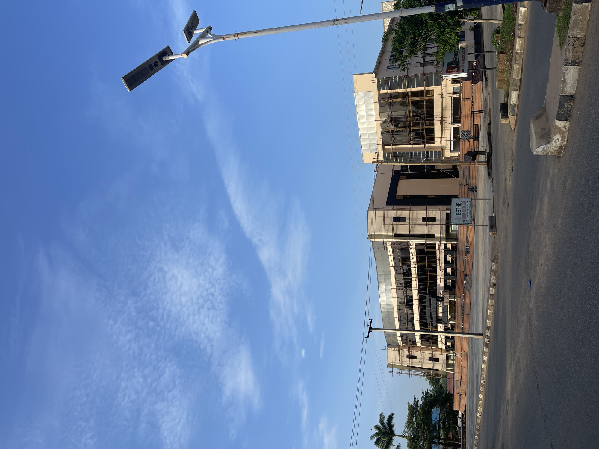 The image depicts a street scene in Bar Jirgi Summa, Nigeria. Two multi-story buildings are under construction, with scaffolding visible on their facades. A street light pole is prominently positioned on the left. Power lines traverse the scene. A paved road and sidewalk occupy the foreground. The sky is blue with scattered clouds. The overall impression is that of a modern, developing urban environment.