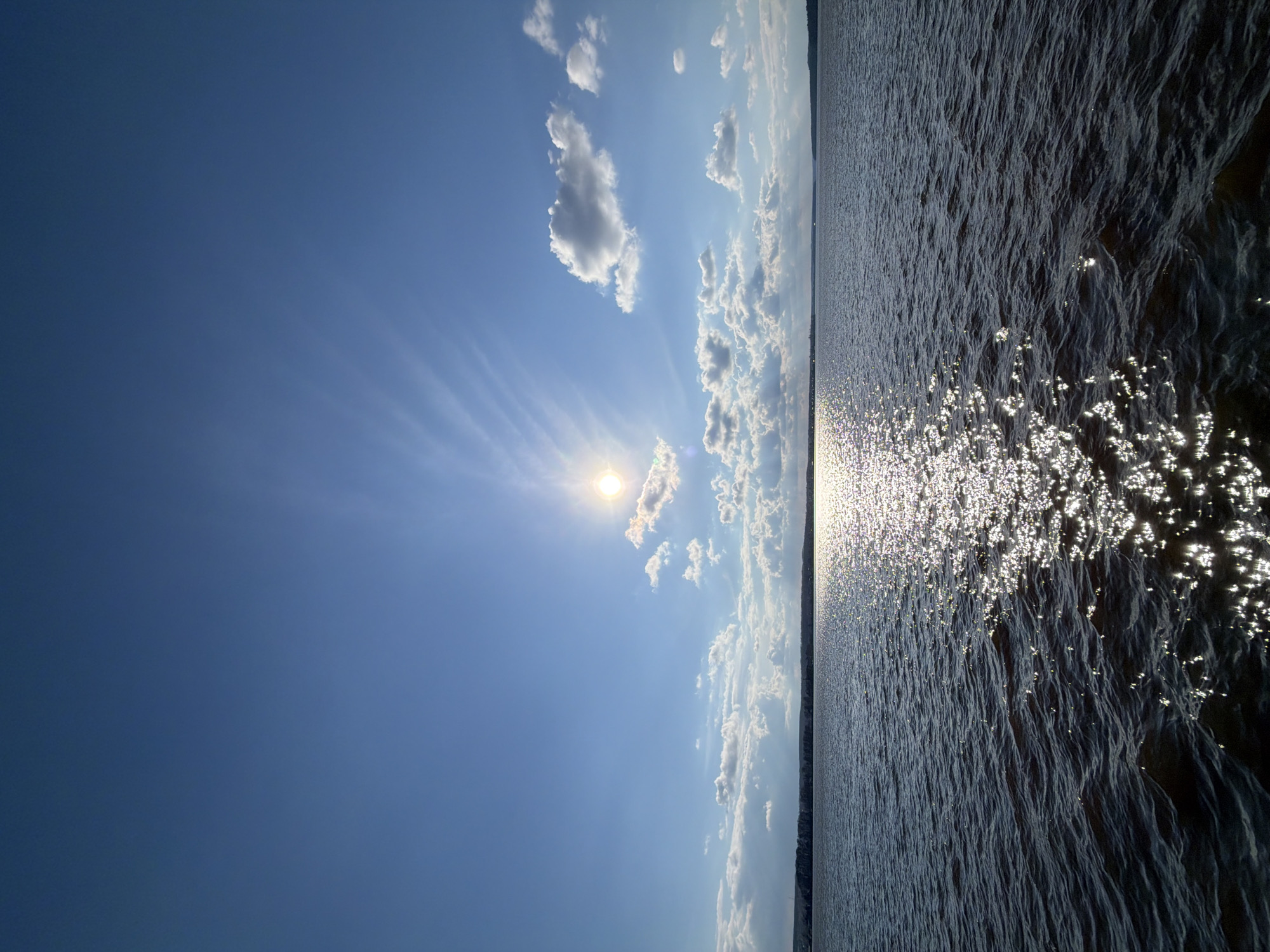 The image presents a serene landscape featuring a vast body of water under a bright blue sky, likely the Votkinsk Reservoir in Votkinsk, Russia. The horizon line extends horizontally across the middle of the frame, dividing the sky from the water.
The upper portion of the image displays a clear blue sky, with the sun prominently positioned on the left side. It emits intense, brilliant rays, creating a distinct sunburst effect. Scattered white cumulus clouds of various sizes are visible, some illuminated by the sunlight, alongside delicate, wispy cirrus clouds in the upper left quadrant.
The lower half of the image is dominated by the expansive water surface, which is textured with ripples and small waves. The strong sunlight reflects intensely off the water, creating numerous glittering, sparkling points that extend across the surface, especially towards the right. A faint, dark line of distant land or a shoreline is discernible along the horizon, indicating the considerable breadth of the water body.
The scene depicts a peaceful daytime moment, suggesting clear and pleasant weather conditions. The sun's position indicates it is likely mid-day or late afternoon. There is no visible human activity or text within the image, emphasizing the natural beauty and tranquility of the setting.