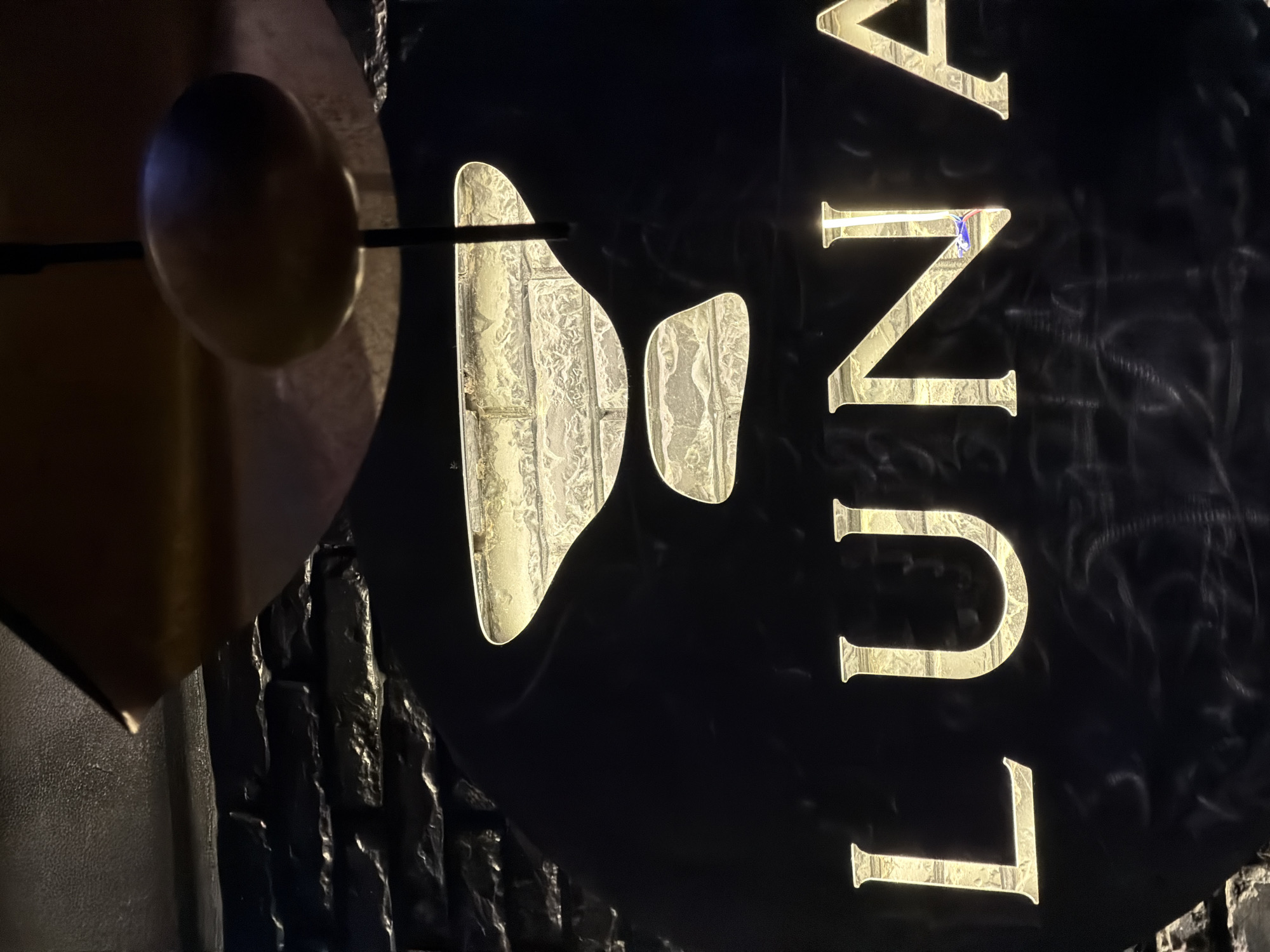 The image shows a sign that reads "LUNA" in illuminated gold letters. The sign is attached to a dark, textured wall that resembles rough brick or stone. To the left of the sign is a dark, round object possibly connected to a rod or fixture. Based on the sign, this could be the Luna Restaurant located in Bishkek City, Kyrgyzstan.