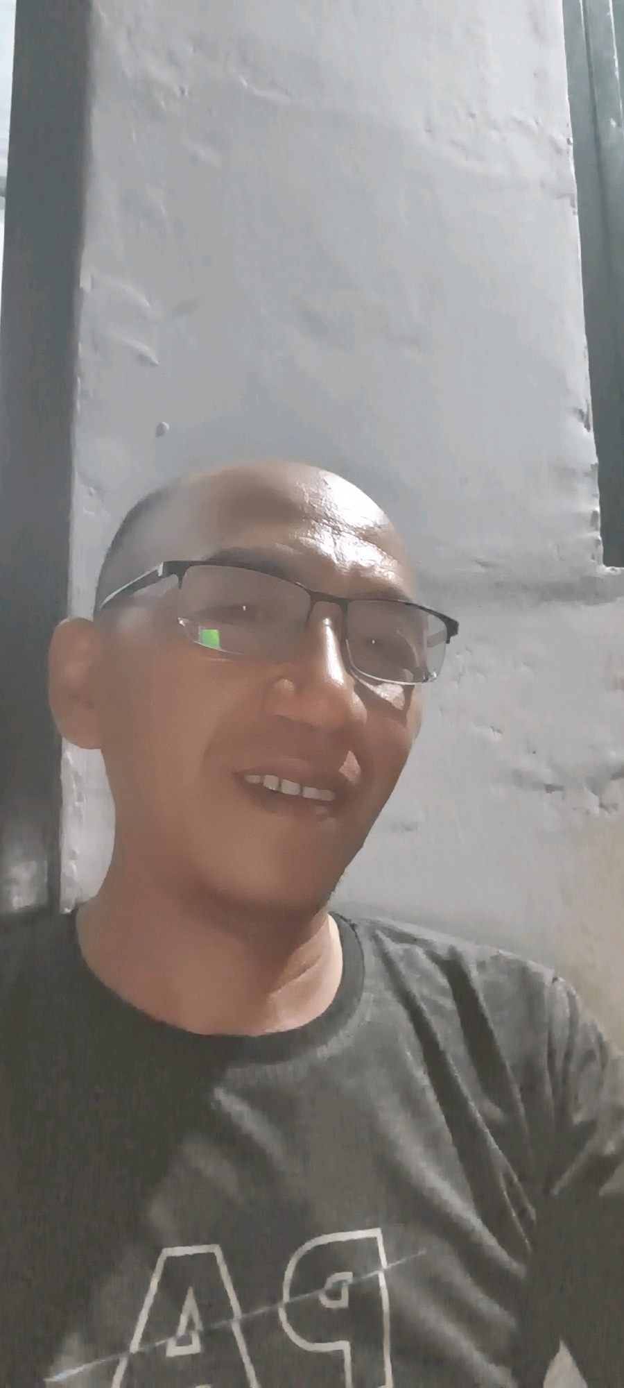 A close-up, head-and-shoulders selfie of a bald, middle-aged man with glasses is shown. He is wearing a dark grey t-shirt with a white graphic on it that appears to say "A9". The man is smiling, and his teeth are visible. He has a light complexion and is wearing black-rimmed glasses. The background is a grey, textured wall, possibly an exterior wall of a building. The lighting suggests it is daytime, and the scene is likely outdoors or in a well-lit indoor space. The image appears to be taken in South Tangerang, Indonesia, given the user's context.