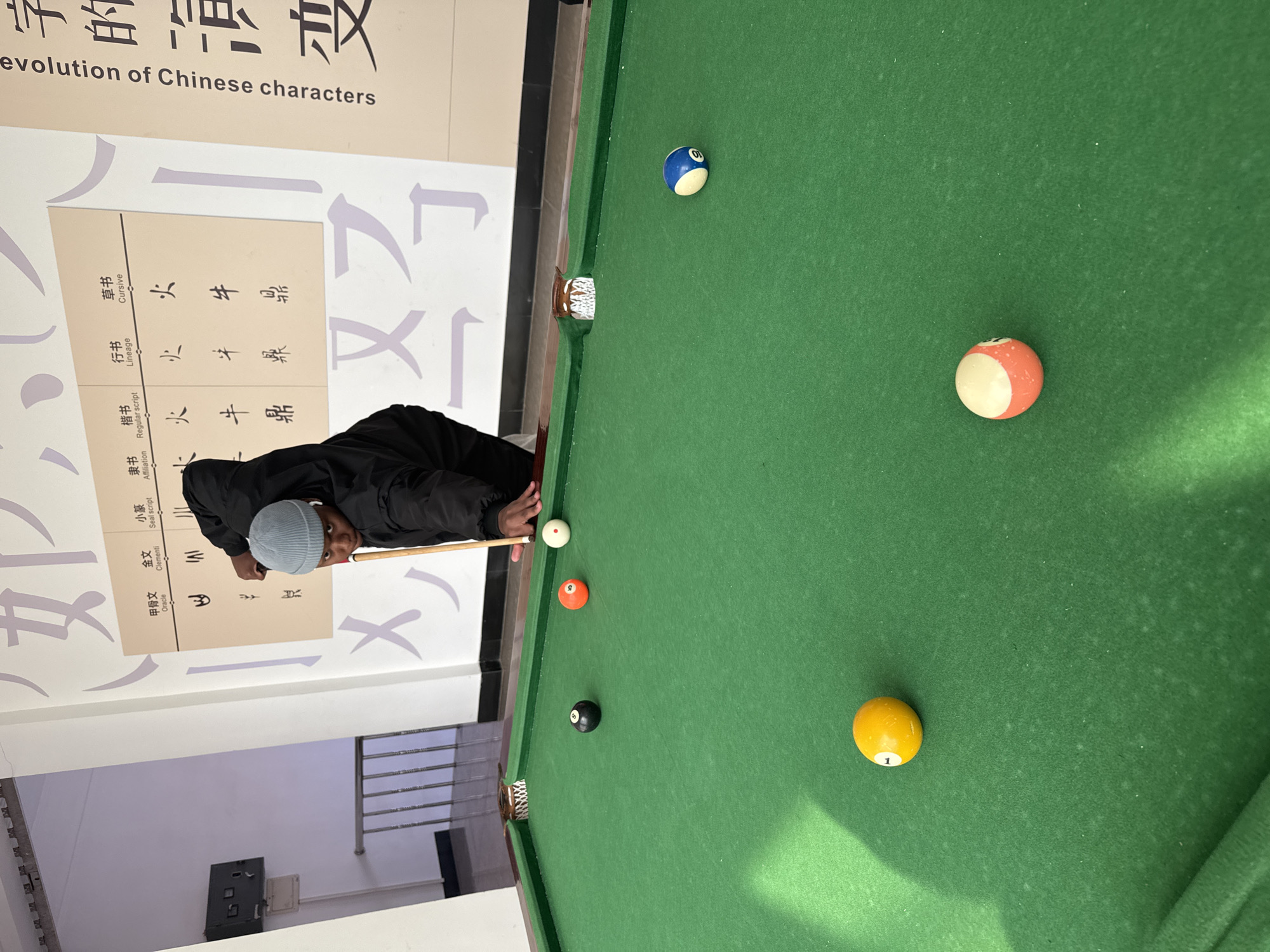 The image depicts a person with dark skin, wearing a black jacket and light blue knit cap, aiming a cue stick at a white cue ball on a green felt pool table. Visible on the table are other billiard balls, including orange, yellow, black, and blue balls, arranged across the surface. The side of the pool table is adjacent to a wall displaying an exhibit about the "evolution of Chinese characters." The exhibit features Chinese writing samples in various styles such as Oracle, Clementi, Seal script, Affiliation, Lineage, and Cursive. Through a doorway, a stairway with metal railings is visible. The specific location is the city of Zhongtun, China.