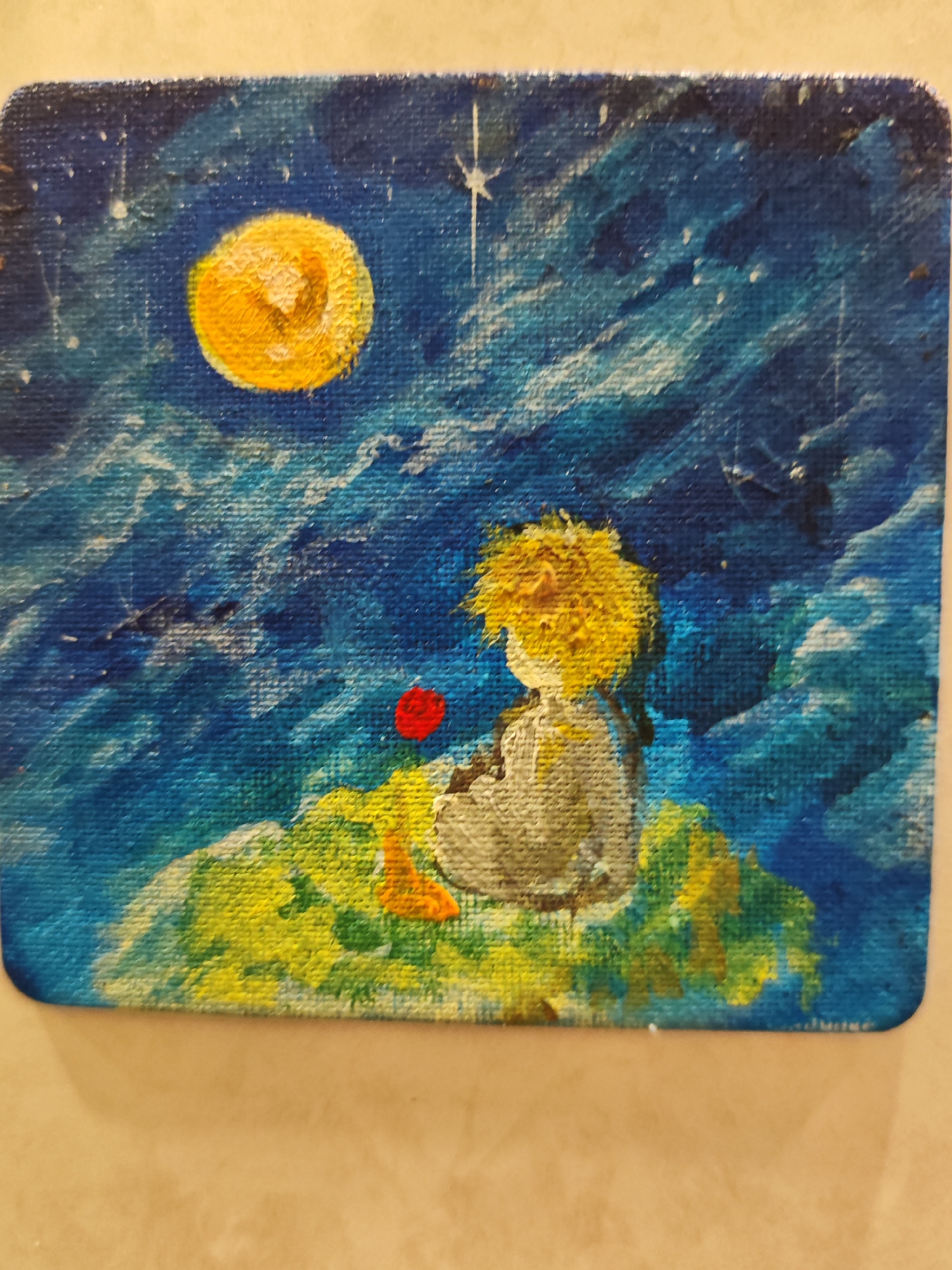The image is a painting of a young boy with blonde hair, seen from the back, sitting on a grassy knoll. He is wearing a light-colored shirt and darker pants. A single red rose is positioned in front of him. Above and to the left, a bright yellow moon hangs in a dark blue, cloudy sky dotted with stars. The overall style is impressionistic, with visible brushstrokes and textured paint. The setting is not identifiable as being in a specific city or location.