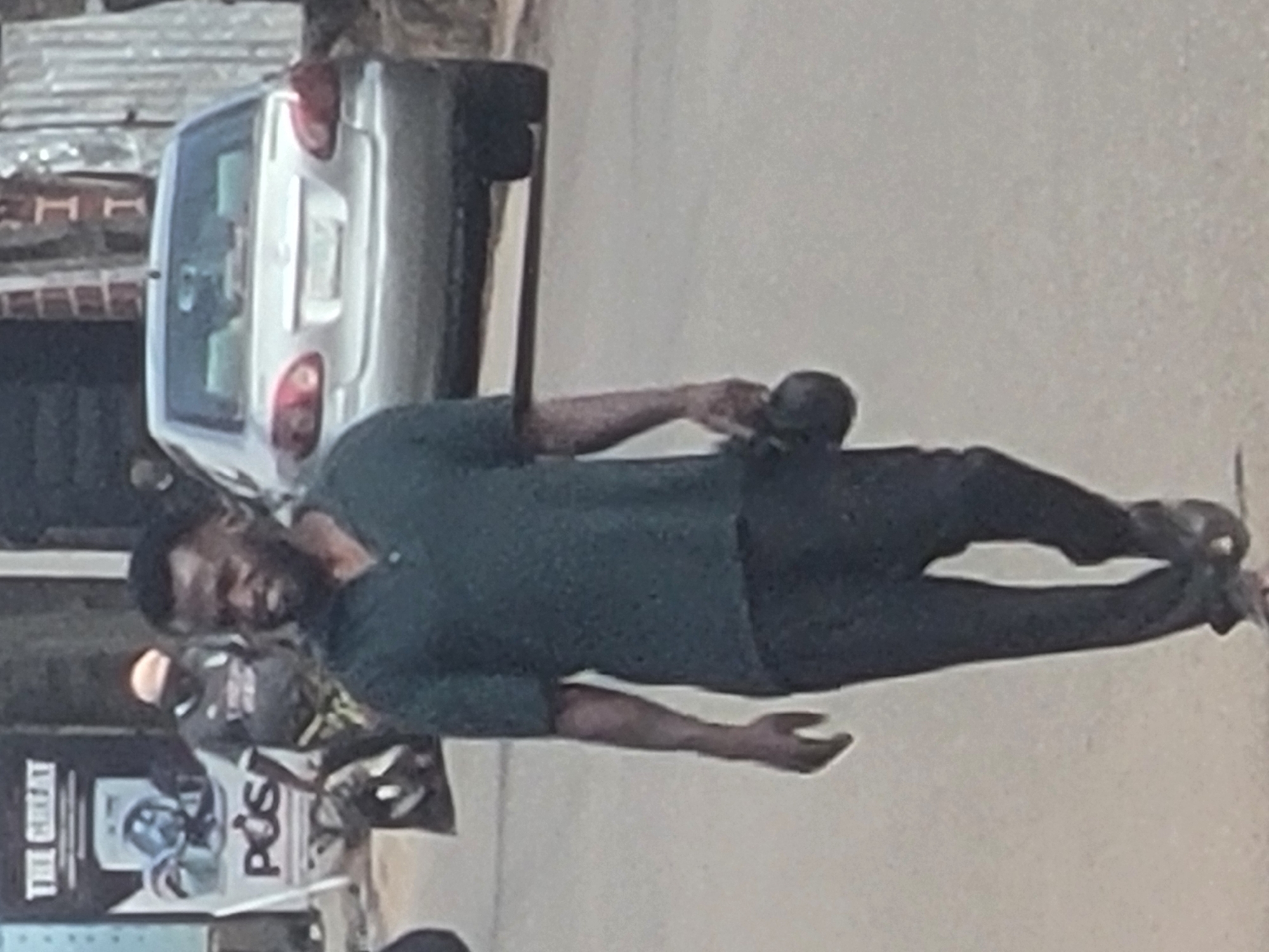 In the city of Jos, Nigeria, a man wearing a dark green short-sleeved shirt and dark pants is lying on a paved surface. He is facing the camera, with a silver sedan parked to his left. Behind him, a poster on a pole or wall is visible. The immediate environment appears to be a street or roadside.
