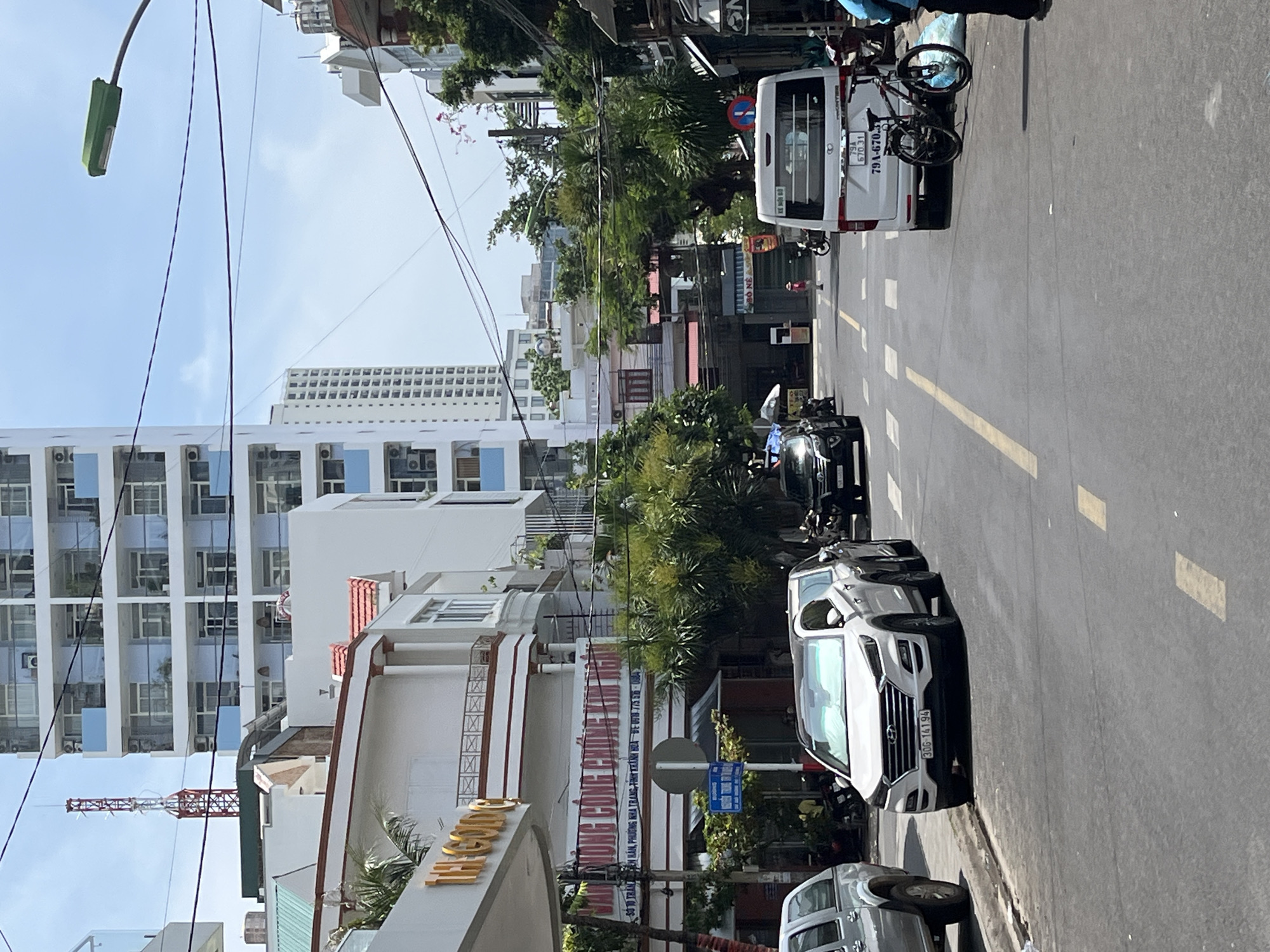 The image shows a street scene in Nha Trang Ward, Vietnam. A white SUV, a white van, and several motorcycles are parked or driving on the asphalt road, which has yellow dividing lines. A bicycle is leaning against the white van. On the left side of the street, a white building with large windows and balconies is visible, along with a green-roofed structure and a red antenna tower in the distance. Further down the street, more buildings of various heights and architectural styles can be seen, interspersed with trees and power lines. The sky is blue with scattered clouds. A few pedestrians are present on the sidewalk and street.