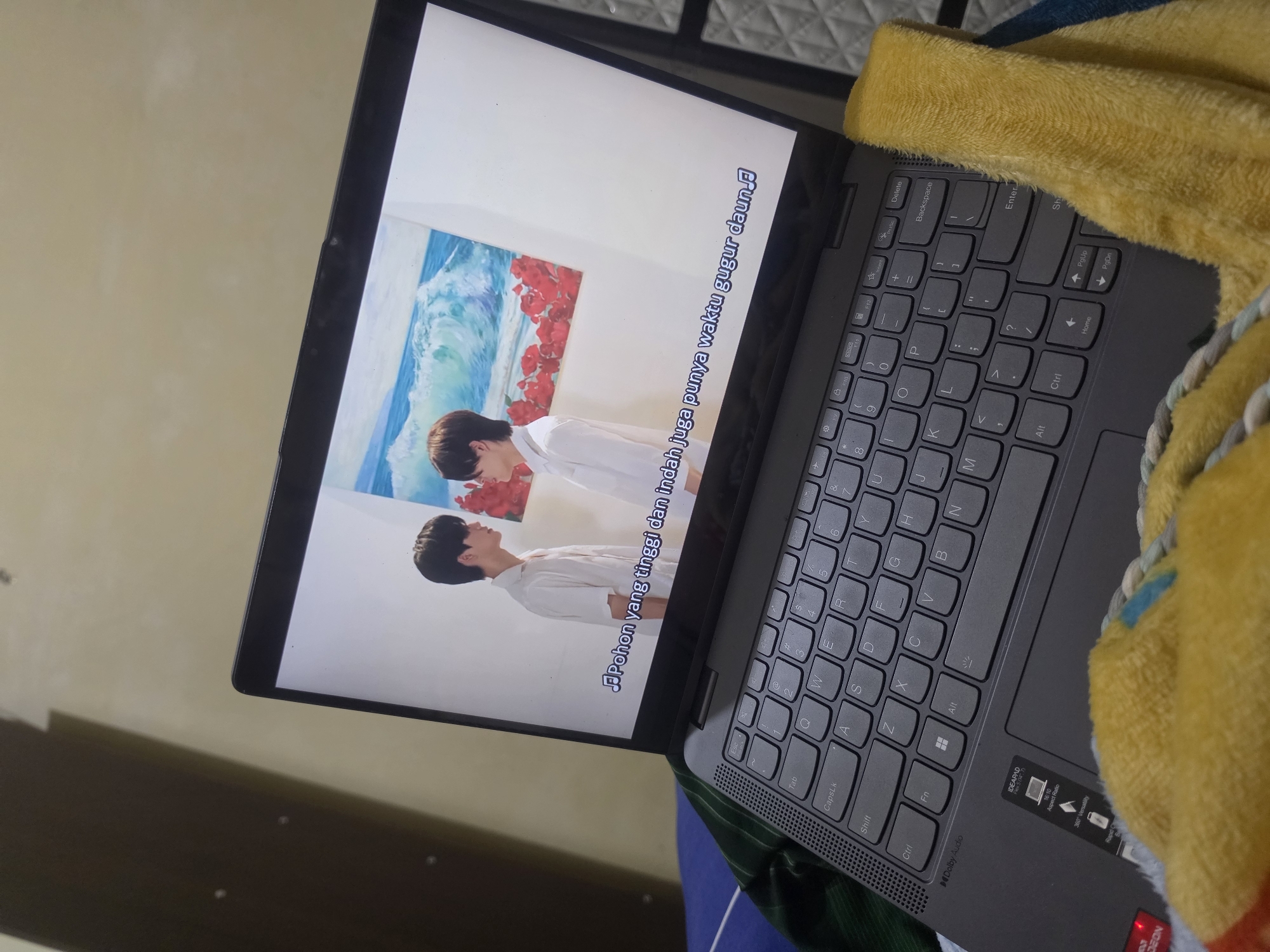 Here's a description of the image:
The image depicts a gray Lenovo laptop sitting on what appears to be a bed. The laptop screen is on and displaying a video, showing two men in front of an artwork depicting an ocean scene with red flowers. Subtitles in Indonesian are displayed on the screen. A yellow towel is partially draped over the keyboard of the laptop. The background is slightly out of focus but appears to be a wall. The image is taken indoors, with a slight diagonal orientation. I do not have information to ascertain the location of this image in Pintomakmur, Indonesia.