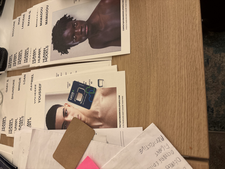 The image shows several headshots from the agency Human Models Paris, stacked on a wooden surface. The headshots display models with their names: Gaby S., Shunsuge, Mingje, Loan, Gabriel, Youssef, Medoune, Ikar, Lancene, Rayan G., Mamadou. A Lyca Mobile SIM card and some handwritten notes on an envelope are also on the stack. The envelope appears to have written in black marker: DEVRED, OCTOBRE EDITION, FIGARET PERSPECTIVE.