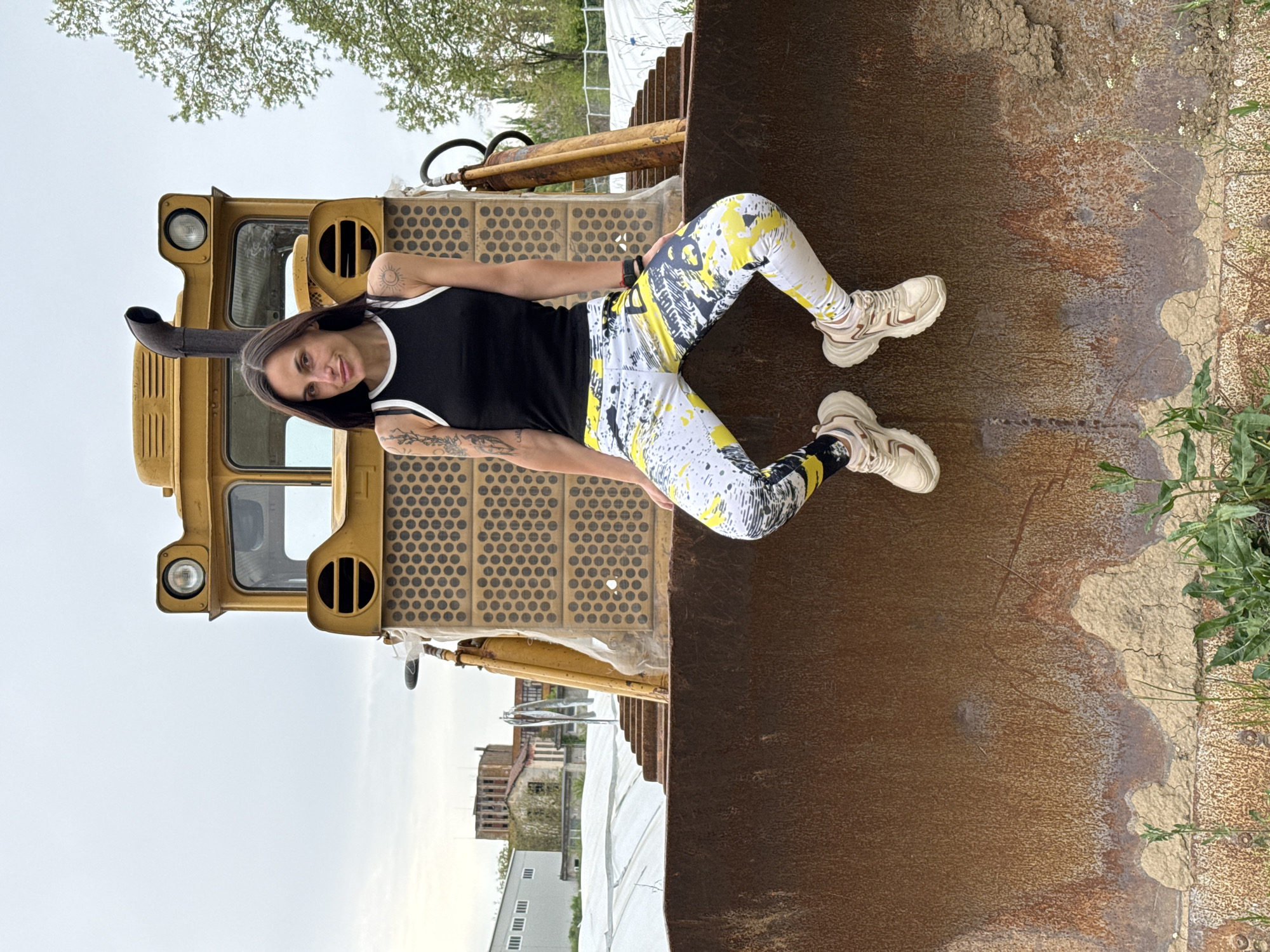A woman with long, dark hair and tattoos is depicted lying on her side on a large, yellow construction vehicle. She wears a black tank top with white trim and white leggings adorned with vibrant yellow, black, and gray abstract splatter patterns, featuring partially obscured stylized text. Her left arm is propped, supporting her upper body, and she looks directly at the camera with a neutral expression. Visible tattoos include a sun-like design on her left shoulder and more abstract patterns on her right forearm.
The setting places her on what appears to be the grated engine cover of a bulldozer or front-end loader, with its cabin, a single headlight, and an exhaust pipe visible on the left side of the frame. To her right, a large, textured, rusty brown surface, possibly part of the vehicle's bucket or an adjacent industrial structure, dominates the lower part of the scene. The background reveals an overcast sky, green tree foliage, and distant buildings, one of which appears to be a derelict or abandoned structure. The scene is illuminated by daylight, suggesting an outdoor setting. The location is Pleven, Bulgaria.