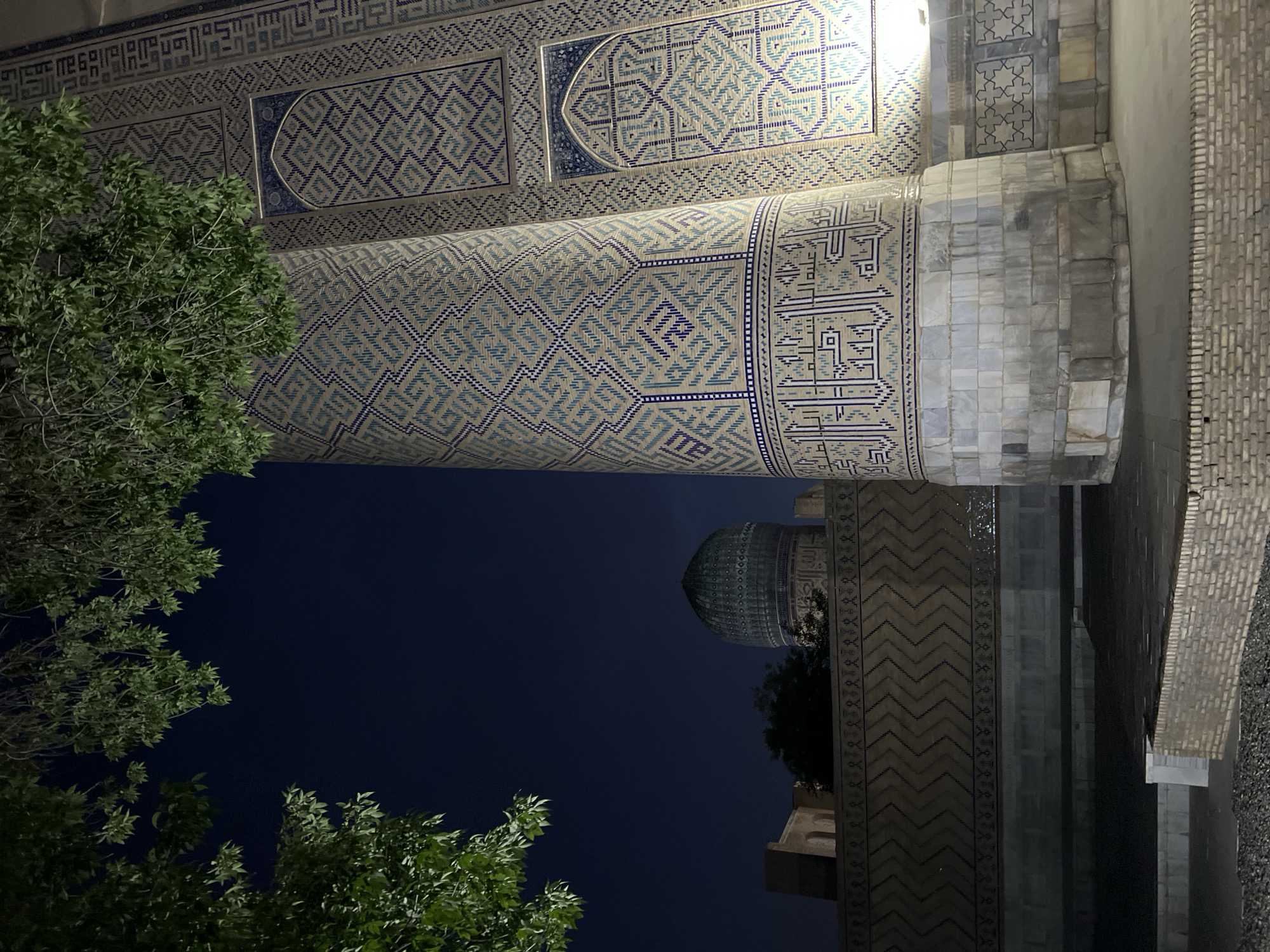 The image captures a nighttime view of a majestic architectural complex, likely a historical religious site in Samarkand, Uzbekistan. The primary subject is the intricately tiled facade of a grand building, featuring geometric patterns in shades of blue and white. Elaborate calligraphy is also visible, adorning sections of the structure.
Dominating the center of the frame is a massive, cylindrical pillar covered in mosaic tiles, which appears to be part of the main building's entrance or support structure. To the right, a section of a brick wall and a paved area suggest an exterior walkway or courtyard. In the background, a distinctive turquoise dome, a hallmark of Islamic architecture, is partially visible against the dark night sky. The dome is surrounded by more decorative tiling.
Green trees with lush foliage frame the left and bottom portions of the image, their leaves illuminated by artificial lighting, creating a stark contrast with the deep blue of the night. The lighting casts a soft glow on the architectural elements, highlighting their detailed artistry.
The time of day is clearly night, evidenced by the dark sky and the artificial illumination of the structures. There are no people visible, suggesting a moment of quiet contemplation or the absence of daytime activity. The overall mood is one of awe and grandeur, emphasizing the historical and cultural significance of the location. The visible text, which is Arabic calligraphy, adds to the rich cultural tapestry of the scene.