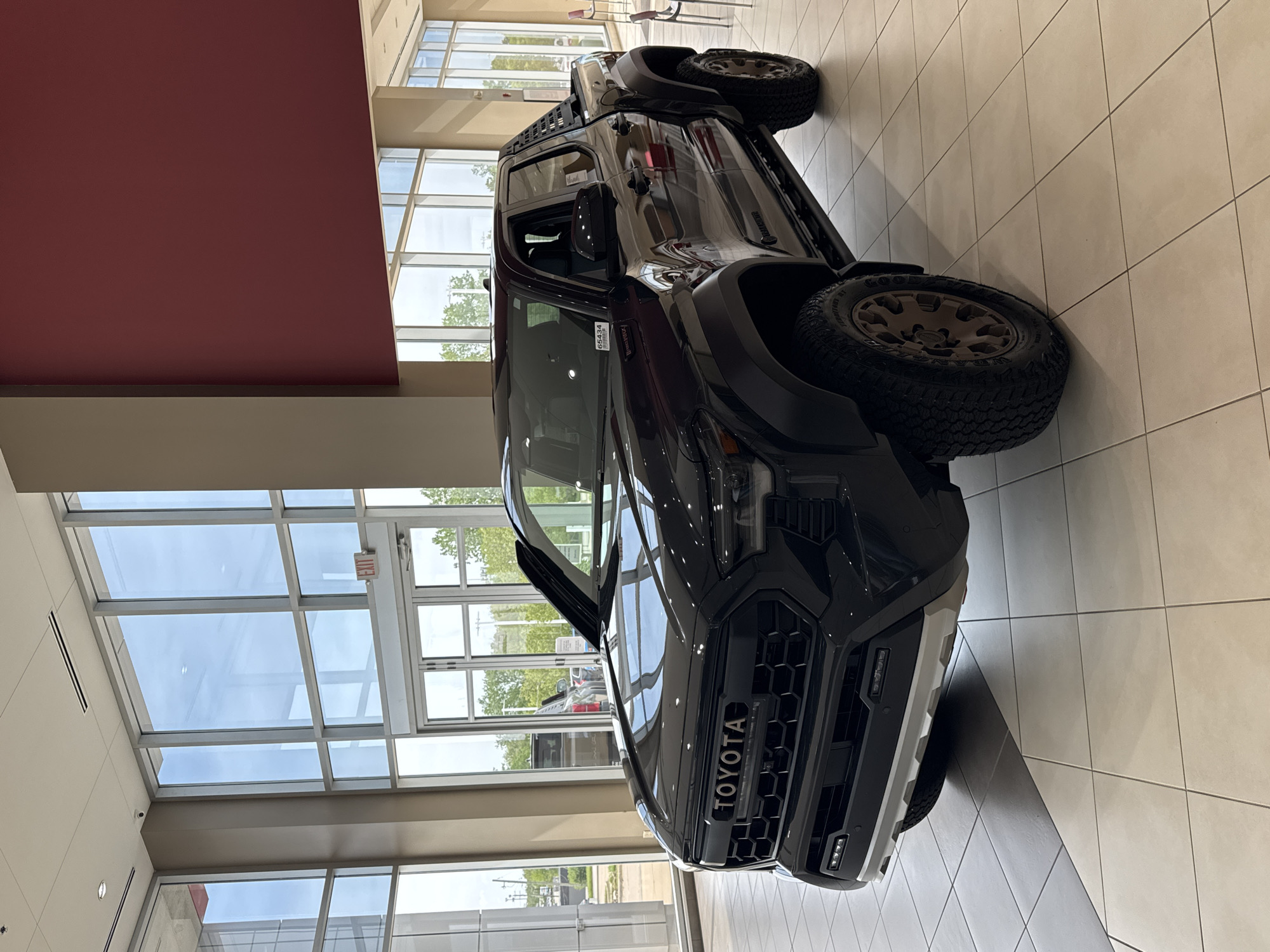 The image depicts a dark-colored Toyota truck, possibly a Tacoma TRD Pro, on display inside a showroom with large windows. The setting appears to be a car dealership with tiled flooring and interior architecture featuring red and beige walls. Natural light floods the scene through the windows. The location is likely in Palatine, United States of America. There are no people visible in the foreground of the image.