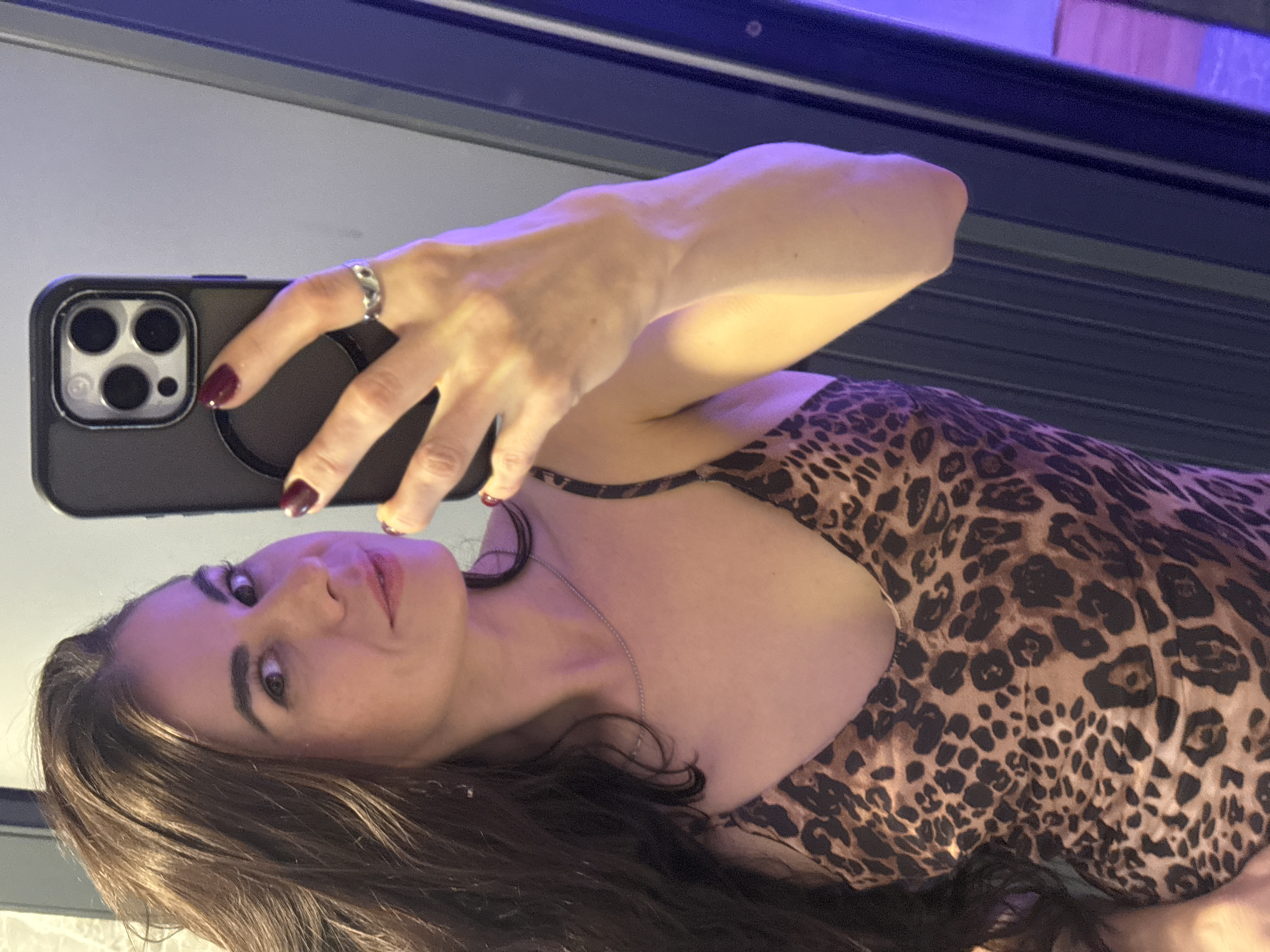 A young woman with long, dark hair is taking a selfie in what appears to be a bathroom. She is wearing a leopard-print dress and a silver ring on her finger. Her nails are painted a dark red color. The background is blurry, but it seems to show a wall with vertical lines. There is no indication that this image was taken in Da Lat, Vietnam.