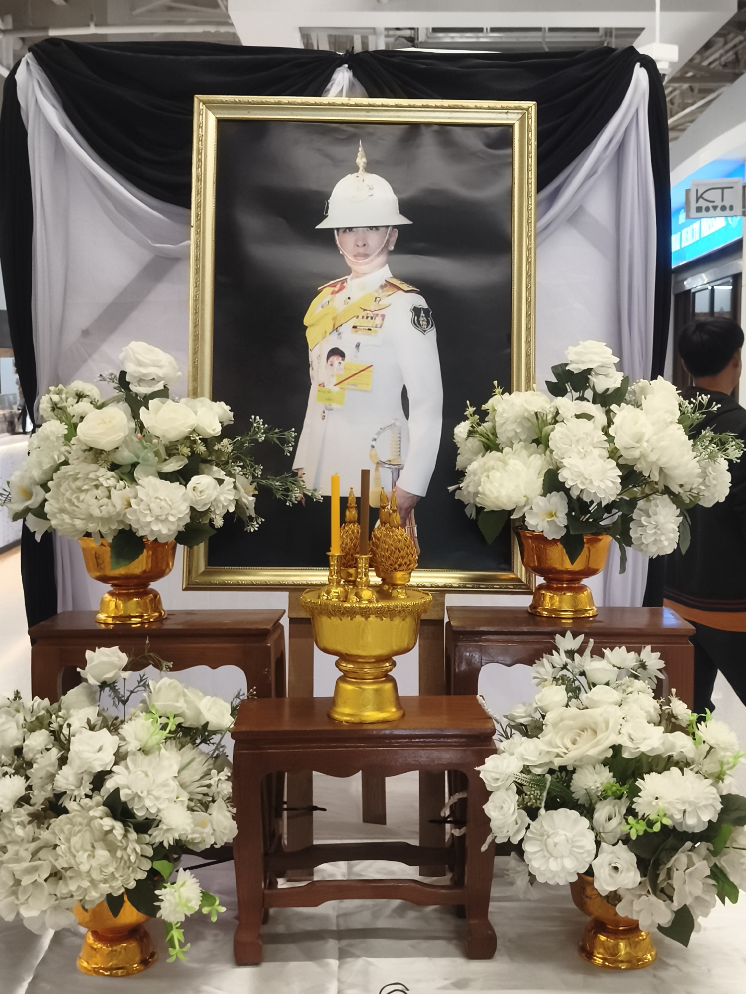 In Baan Lamai, Thailand, a formal memorial setting is depicted, centered around a large framed portrait of a person in military attire, wearing a white helmet and sash. The portrait is framed in gold and hangs against a backdrop of black and white draped fabric. In front of the portrait, on wooden stands, are arrangements of white flowers in ornate gold vases, and golden decorative items, including candles and what appear to be ornate candle holders or incense burners. The scene is further adorned with additional white floral arrangements on smaller wooden tables. A person is visible in the background on the right, facing away from the camera. The overall atmosphere is somber and respectful.
