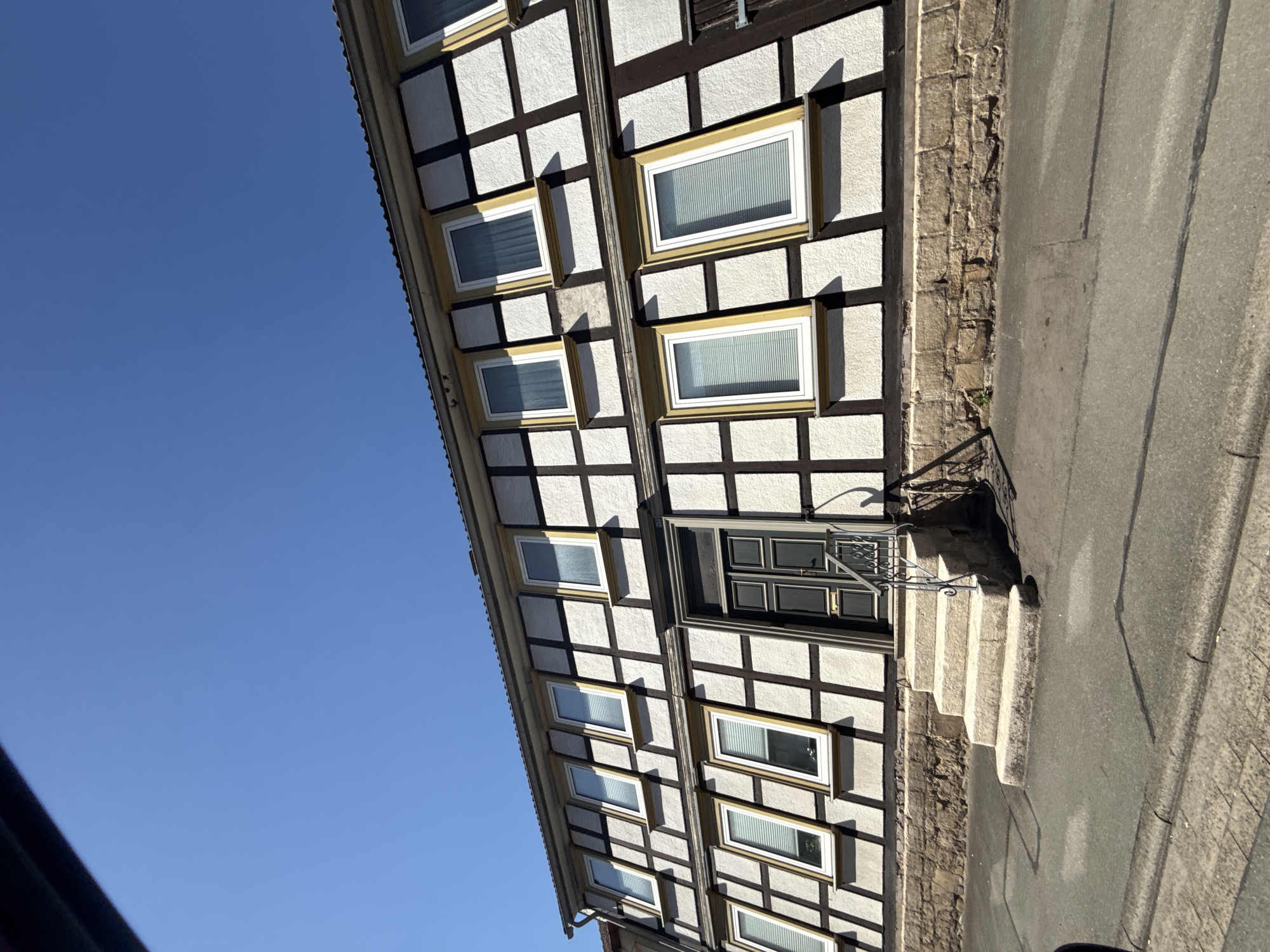 A low-angle, exterior, eye-level shot captures the facade of a two-story half-timbered house under a clear blue sky. The house, located at Hauptstraße 70 in Bad Lauterberg im Harz, Germany, features a distinctive black and white timber frame construction with white infill. The first story has a foundation of rough-hewn stones. A set of three stone steps with a black wrought-iron railing leads up to a dark gray double door. Numerous windows with white frames and beige sills are visible on both floors, many with slatted blinds. The house has a dark tiled roof. The foreground consists of an asphalt street and sidewalk. There are no people or animals visible in the image.