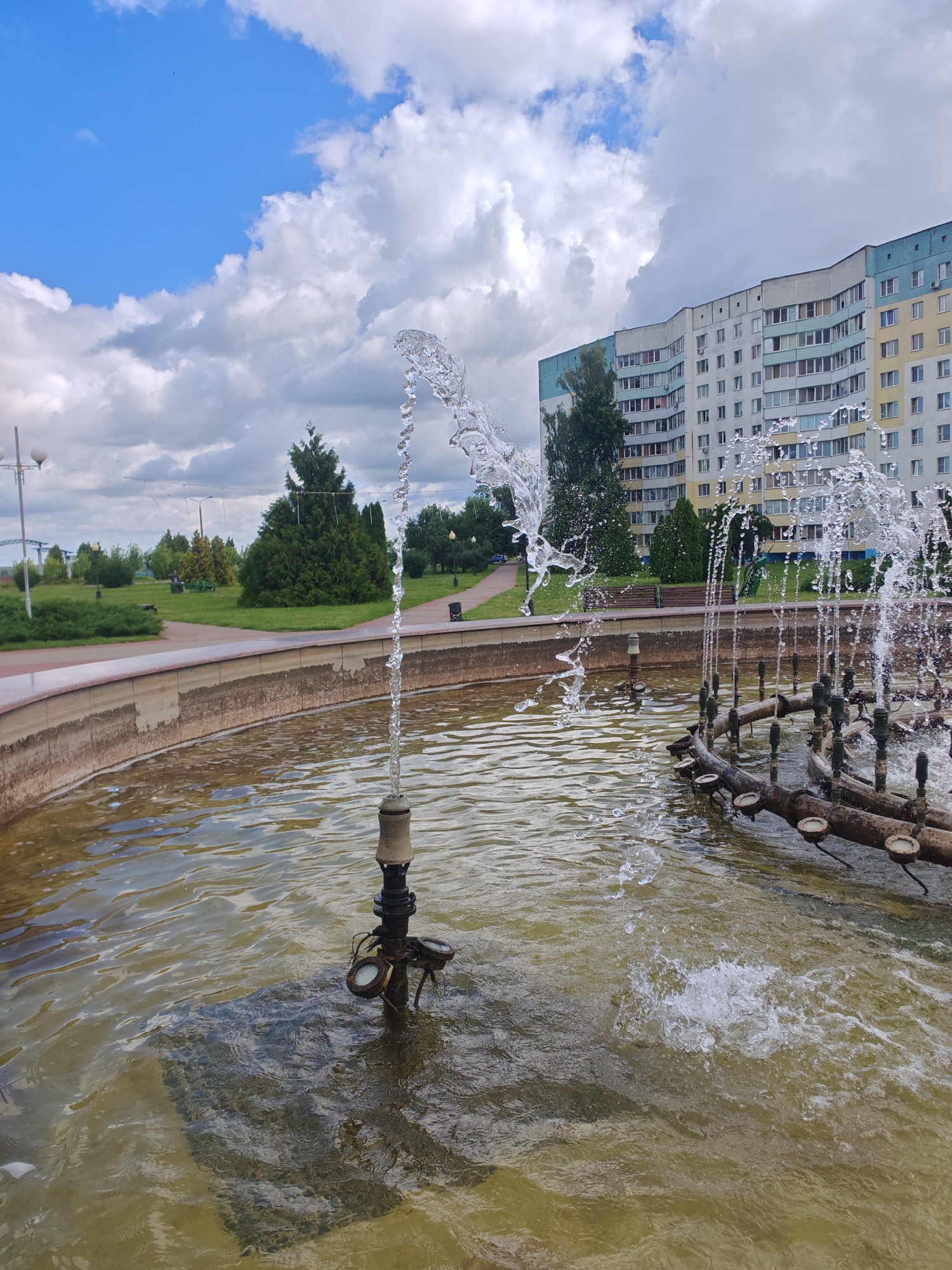 The image shows a fountain with water spraying upwards in a park-like setting. In the background, a large, multi-story apartment building is visible, typical of Soviet-era architecture. The building has a beige and light blue facade with numerous windows. Trees and green grass surround the fountain, and a paved pathway can be seen leading away from it. The sky is partly cloudy, with a mix of blue sky and white clouds. This scene is likely located in the city of Rechytsa, Belarus, a typical urban environment in the region. There are no people visible in the immediate foreground of the fountain, though there might be some in the distance on benches. The fountain itself appears to be made of concrete and metal pipes.