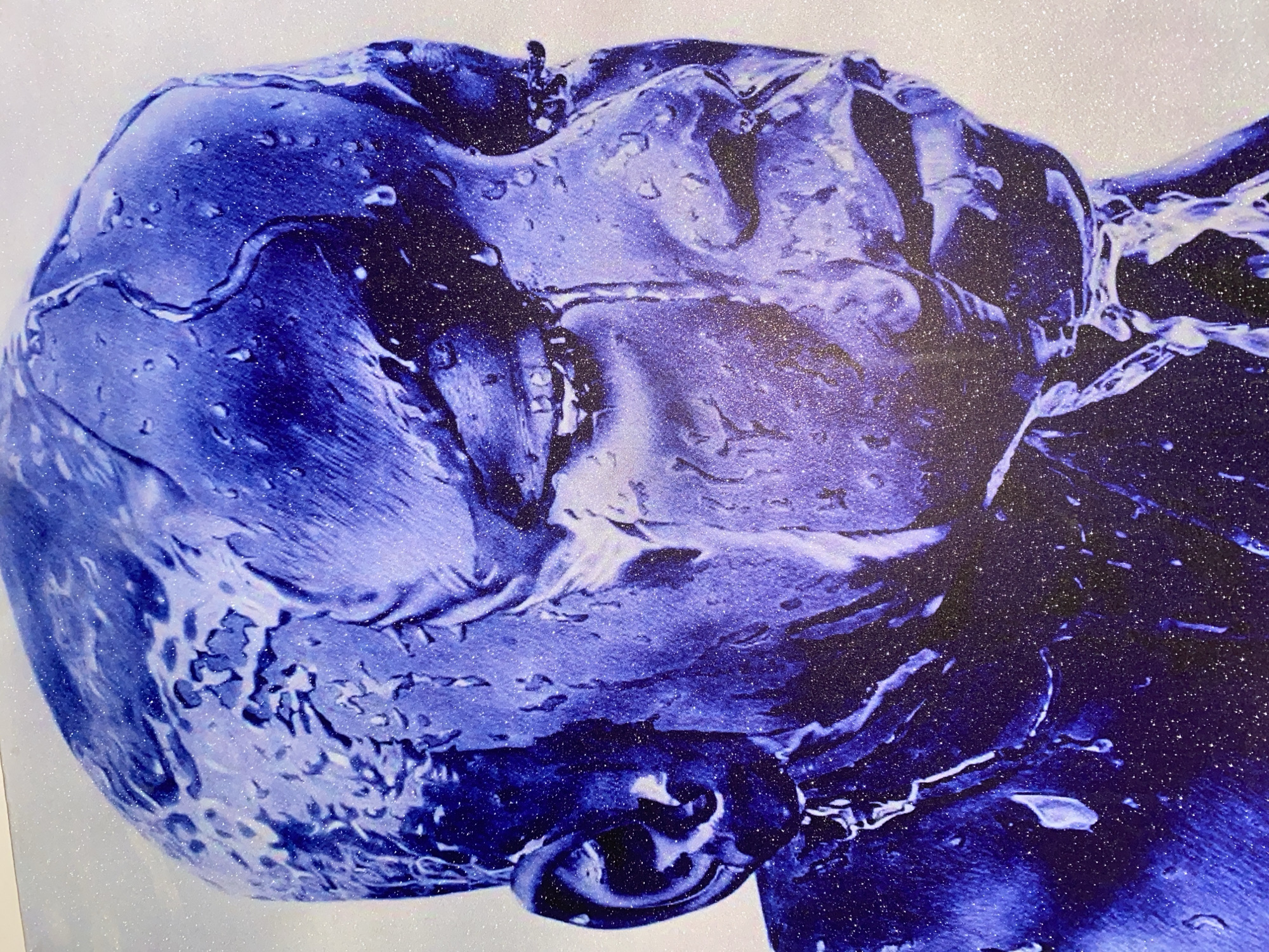 This is a close-up, abstract artwork featuring a depiction of a human head covered in a textured, glossy blue substance that resembles water or paint. The surface is highly reflective, with highlights and shadows that create a sense of depth and form. Tiny white specks, resembling glitter or fine spray, are scattered across the entire surface, adding a shimmering quality. The overall impression is one of fluidity and distortion, with the features of the face subtly suggested rather than clearly defined. The background is a plain, textured white. The artwork evokes a sense of immersion or being enveloped, with the blue material flowing over and obscuring the underlying form. There is no visible text or discernible environmental context. The location context of Giginyu, Nigeria, does not provide any specific visual cues within the image itself.