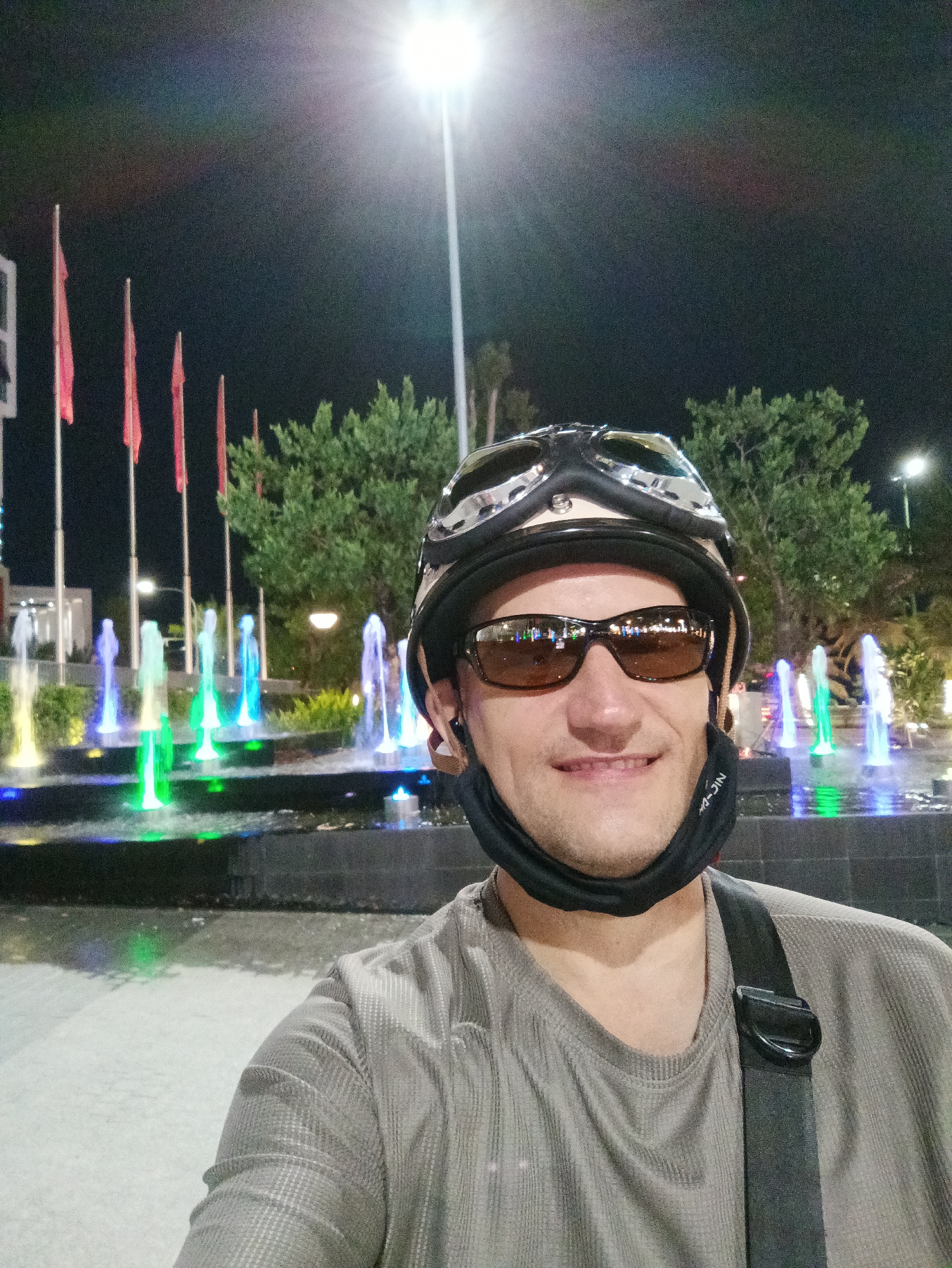 This is a selfie taken at night in Nha Trang Ward, Vietnam. The subject is a person wearing a helmet with goggles, sunglasses, and a black mask pulled down around their neck. They are smiling at the camera. Behind them, a fountain is lit with colorful lights of green, blue, and yellow, creating a vibrant spectacle. Several red flags on tall poles are visible in the background, along with lush green trees. The overall setting appears to be a public space, possibly a park or plaza, illuminated by bright overhead lighting. The time of day is clearly nighttime, and the weather seems to be clear, allowing for the enjoyment of the outdoor fountain display. The image conveys a sense of enjoyment and exploration, suggesting the person is on an outing or adventure in the city. There is visible text on the black mask: "NIC-JIN".