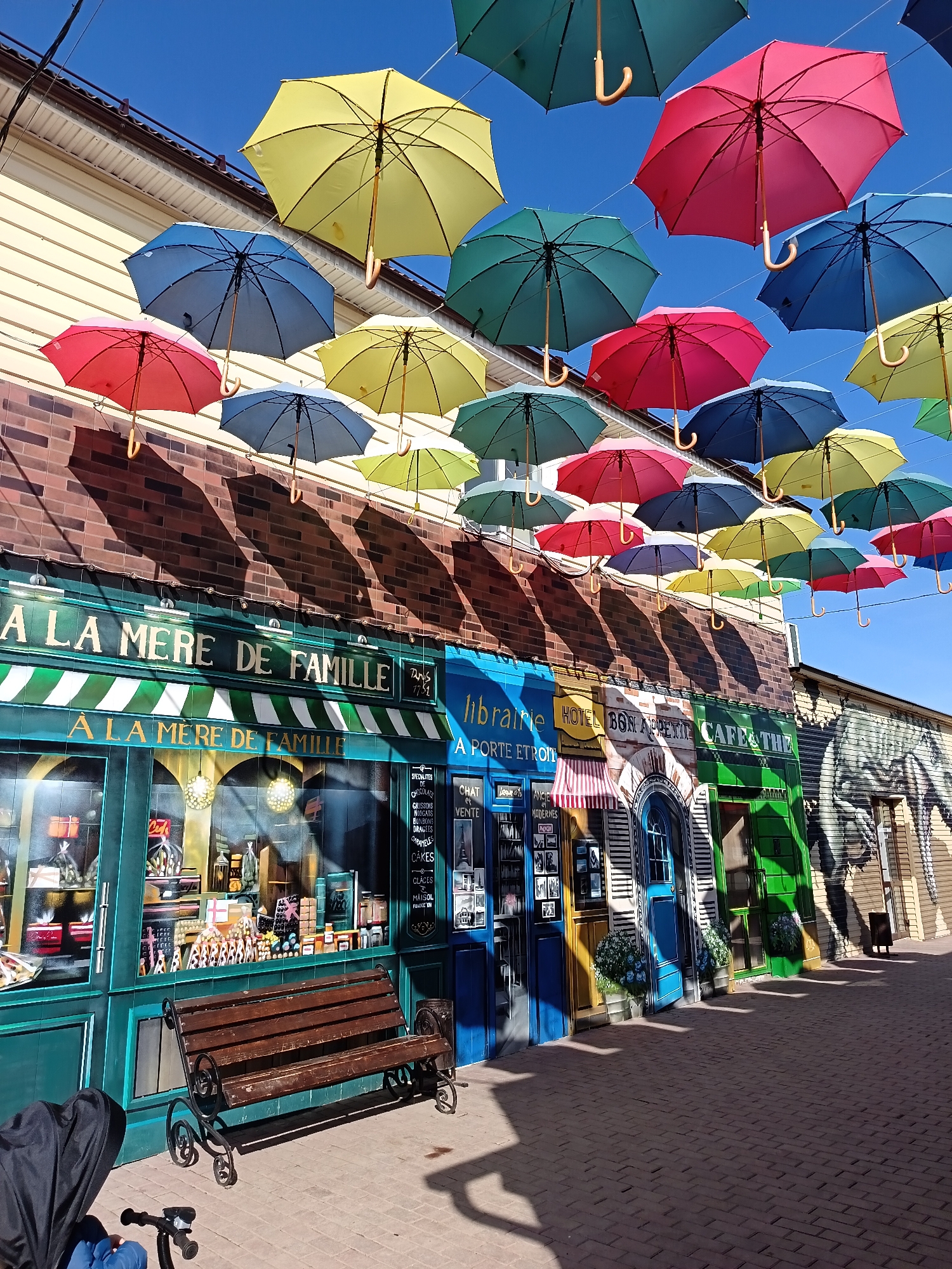The image captures a vibrant street scene in the city of Osa, Russia. Overhead, a colorful display of numerous suspended umbrellas creates a canopy. Below, the facades of various businesses are painted to resemble storefronts, including "À LA MERE DE FAMILLE," "Libraire," and "Hotel Bon Appetit Cafe & The," complete with painted doors and windows.
A bench is placed in front of one of the storefronts, adding a touch of realism to the scene. A baby stroller partially visible on the lower left corner, suggests the presence of people, although none are directly visible. The scene is bathed in bright sunlight, casting shadows of the umbrellas onto the facades and the brick-paved street. In the background, a mural of an animal is painted on a building.