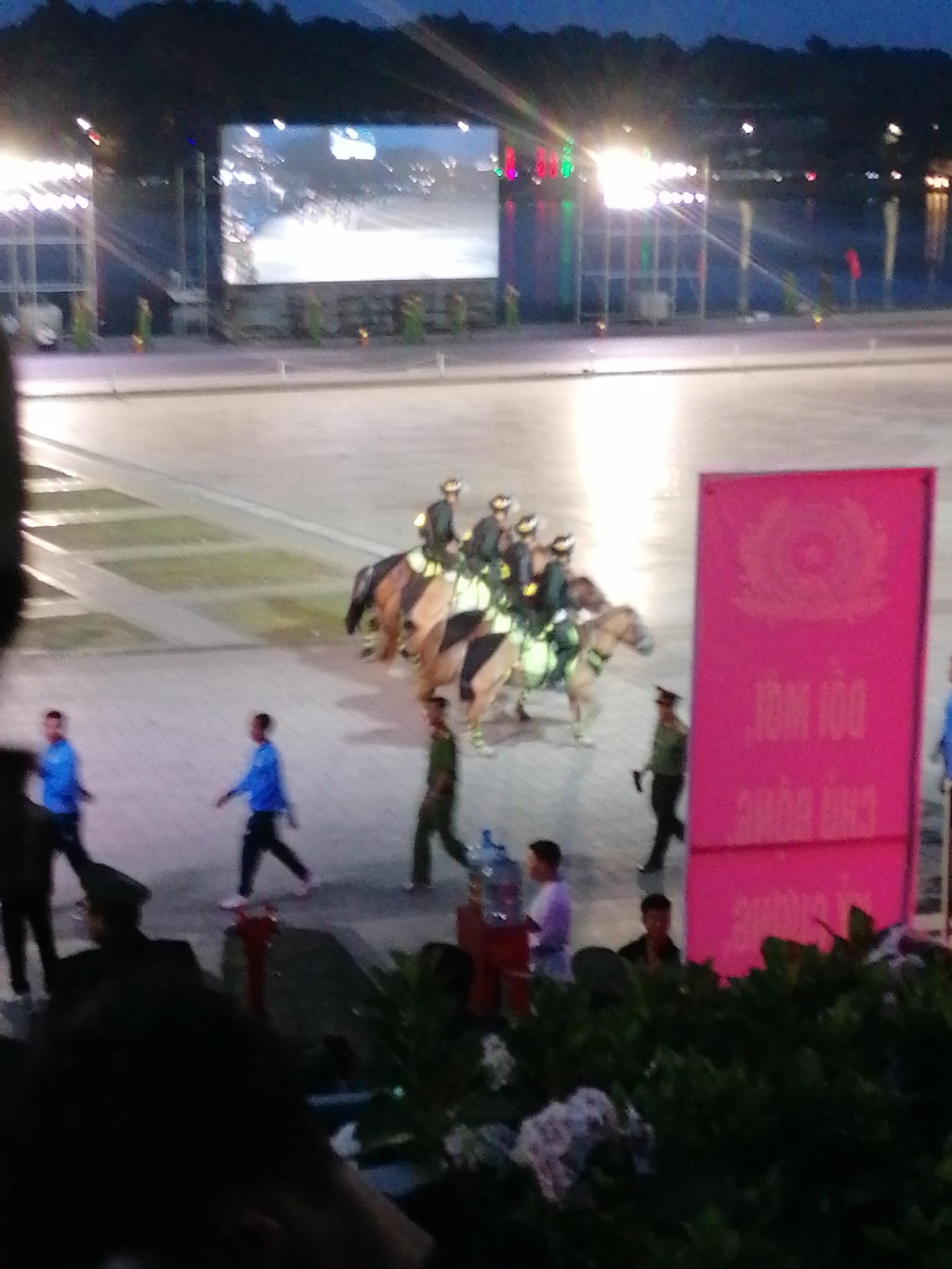 The image depicts an outdoor evening event in Da Lat, Vietnam. In the background, a large screen displays an illuminated image, likely a video or still, set against a backdrop of dark trees. To the left of the screen, a structured lighting rig is visible.
The foreground features a paved area where several individuals are present. A group of people on horseback, wearing military-style uniforms and helmets, are positioned towards the right side of the frame. They are adorned with light-colored vests, possibly reflective gear. In front of the horses, a man in a green uniform with a cap is walking.
Further to the left, two men in bright blue shirts and dark pants are walking. Their stride suggests they are moving with purpose. Other figures are sparsely scattered throughout the scene. To the far right, a large, vibrant pink sign displays text and an emblem, possibly a government or official notice. In the lower right quadrant, lush green foliage partially obscures the scene, with hints of decorative elements or smaller structures within the greenery. The overall lighting suggests twilight or early evening.