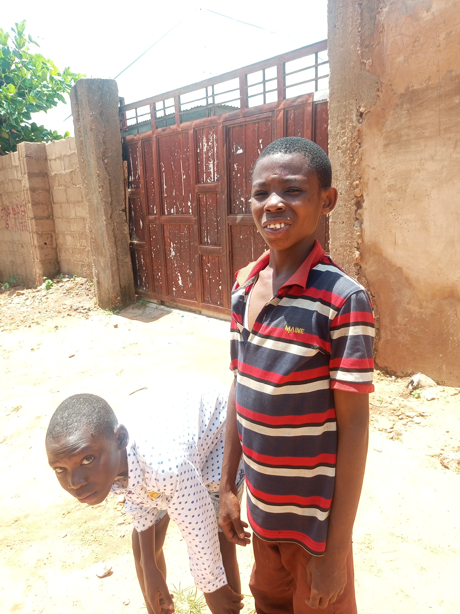 The image depicts two young boys in an outdoor environment. The boy on the right is standing and looking towards the camera, wearing a striped polo shirt with "MAINE" written on it, and brown shorts. He has a wide grin, showing his teeth. The boy on the left is bent over, looking up at the camera with a serious expression. He is wearing a white shirt with blue polka dots.
The background features a large, weathered, reddish-brown metal gate with vertical bars and horizontal supports. To the left of the gate, there is a brick wall and a large, rough concrete pillar. Further to the left, there are green leafy branches of a tree. The ground is sandy and dusty, with some small stones and patches of dry grass visible. The overall impression is of a sunny day in a somewhat rural or undeveloped area, possibly a compound or yard. The location is identified as Tempure, Nigeria.