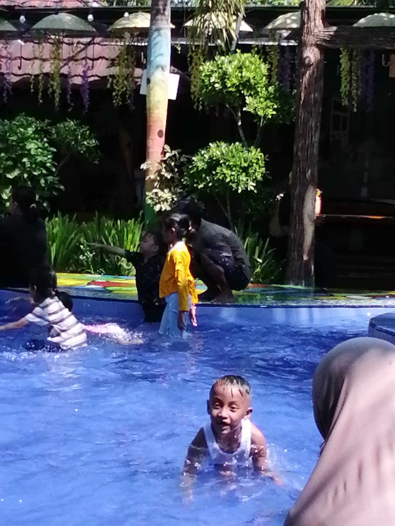 In the city of Tegalwangi, Indonesia, a group of children and adults are enjoying a day at a swimming pool. The pool is filled with clear blue water, and the surrounding area is lush with green vegetation. In the foreground, a young boy is splashing in the water, a wide smile on his face. He is wearing a white tank top. To his right, a person wearing a hijab is partially visible. In the mid-ground, a child in a striped shirt is playing in the water, creating ripples. Another child in a yellow shirt and light blue shorts is standing near the edge of the pool. In the background, several adults are visible, some standing and some crouching. The environment is decorated with hanging plants and umbrellas, suggesting it's an outdoor recreational area. The scene captures a moment of fun and relaxation in a tropical setting.
