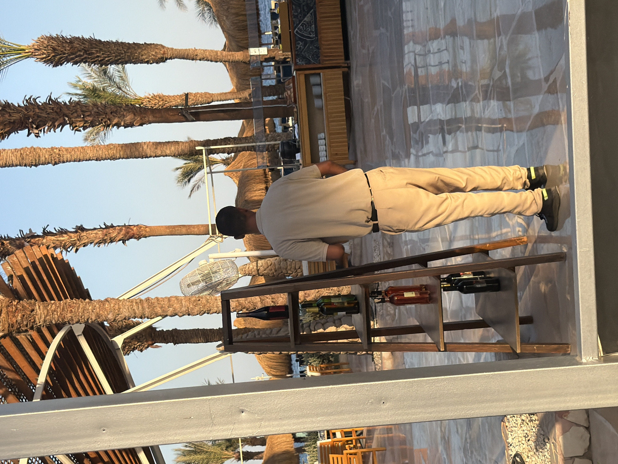 The image depicts a man, possibly a hotel employee, wearing a light-colored polo shirt and tan pants, standing in front of a wooden display shelf holding several bottles. The shelf is located outdoors, with palm trees and thatched-roof structures in the background. The ground appears to be paved with large tiles, reflecting the surroundings. In the distance, a body of water, likely the Red Sea, is visible, and beyond that, the sky is a clear blue. The overall setting suggests a resort or outdoor dining area in Sharm El Sheikh, Egypt, a popular tourist destination known for its beaches and water activities.