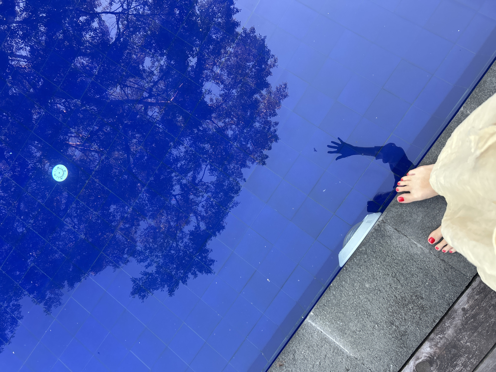 A top-down shot depicts a swimming pool with reflective blue tiles, the surface mirroring a tree and the shadow of a person's hand. The edge of the pool is visible, made of gray stone. A person stands on the pool's edge, their feet with red nail polish and part of a light-colored dress are in the frame. The location is Kediri, Indonesia.