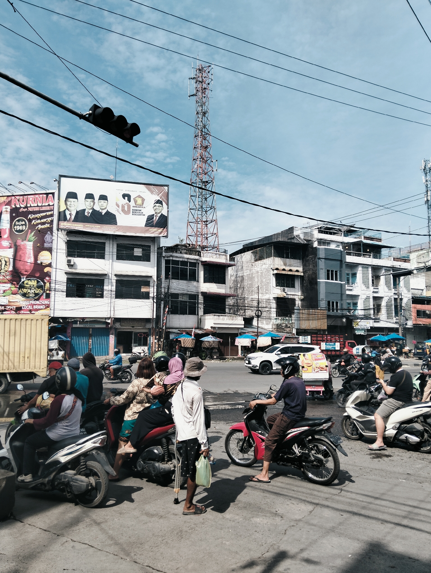 The image depicts a busy street scene in the City of Medan, Indonesia, under a bright, cloudy sky. A prominent telecommunications tower stands tall in the background, with numerous power lines crisscrossing the sky. Several multi-story buildings line the street, some appearing older and weathered, others more modern.
The foreground is dominated by a busy intersection where various people on motorcycles are waiting, suggesting traffic or a pause in movement. A man on a red motorcycle in the center is looking to his left, while another man on a white motorcycle to his right is looking down. A man with a walking stick and a plastic bag stands to the right of these motorcyclists. Several other individuals on motorcycles are visible, either waiting or in motion.
On the left side of the image, a large billboard displays political figures and campaign slogans. Adjacent to it is a yellow truck. In the mid-ground, a white car is visible, and other vehicles and motorcycles are scattered throughout the scene. Blue and white awnings or stalls are set up along the side of the road, indicating commercial activity. The overall impression is of a typical, vibrant urban environment in Indonesia.