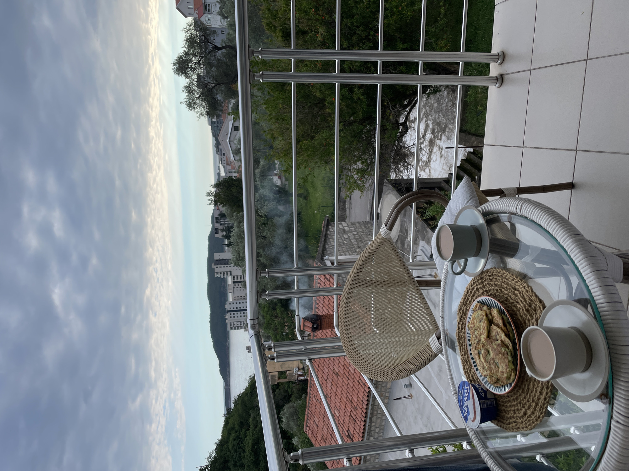 The image shows a balcony scene in Rafailovići, Montenegro. The balcony has a silver railing and white tile flooring. A round glass-topped table with a white wicker base is placed on the balcony. On the table are two cups of coffee, a plate of food resembling pancakes or fritters, and a small container of yogurt. A woven placemat sits under the food items. A beige wicker chair is positioned near the table.
In the background, the view from the balcony includes buildings, trees, and a body of water. Smoke is visible rising from a building in the middle ground. The sky is overcast with clouds.