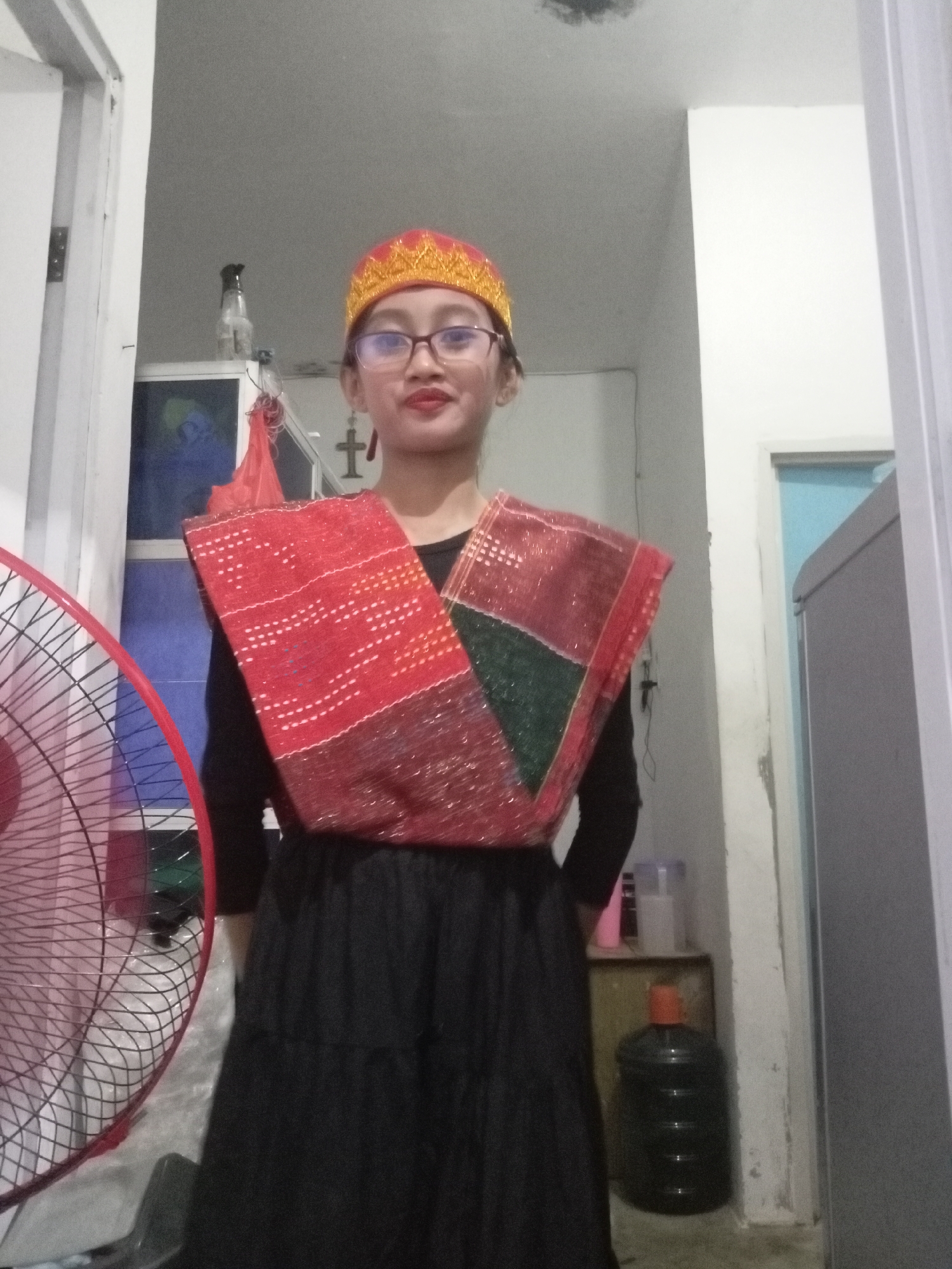 A young person wearing a red and gold headdress, glasses, and a black long-sleeved shirt stands in front of a patterned fabric draped over their shoulders. The fabric has red and green sections with intricate white and gold embroidery. They are wearing a long black skirt. To the left of the frame, a red electric fan is visible. The background appears to be an indoor residential setting, possibly a hallway or a room with a door and a closet. A water dispenser is visible in the background to the right. The lighting suggests it is daytime or an interior with artificial light. The scene does not provide specific clues to pinpoint the exact location within Batam, Indonesia, beyond it being a residential interior.