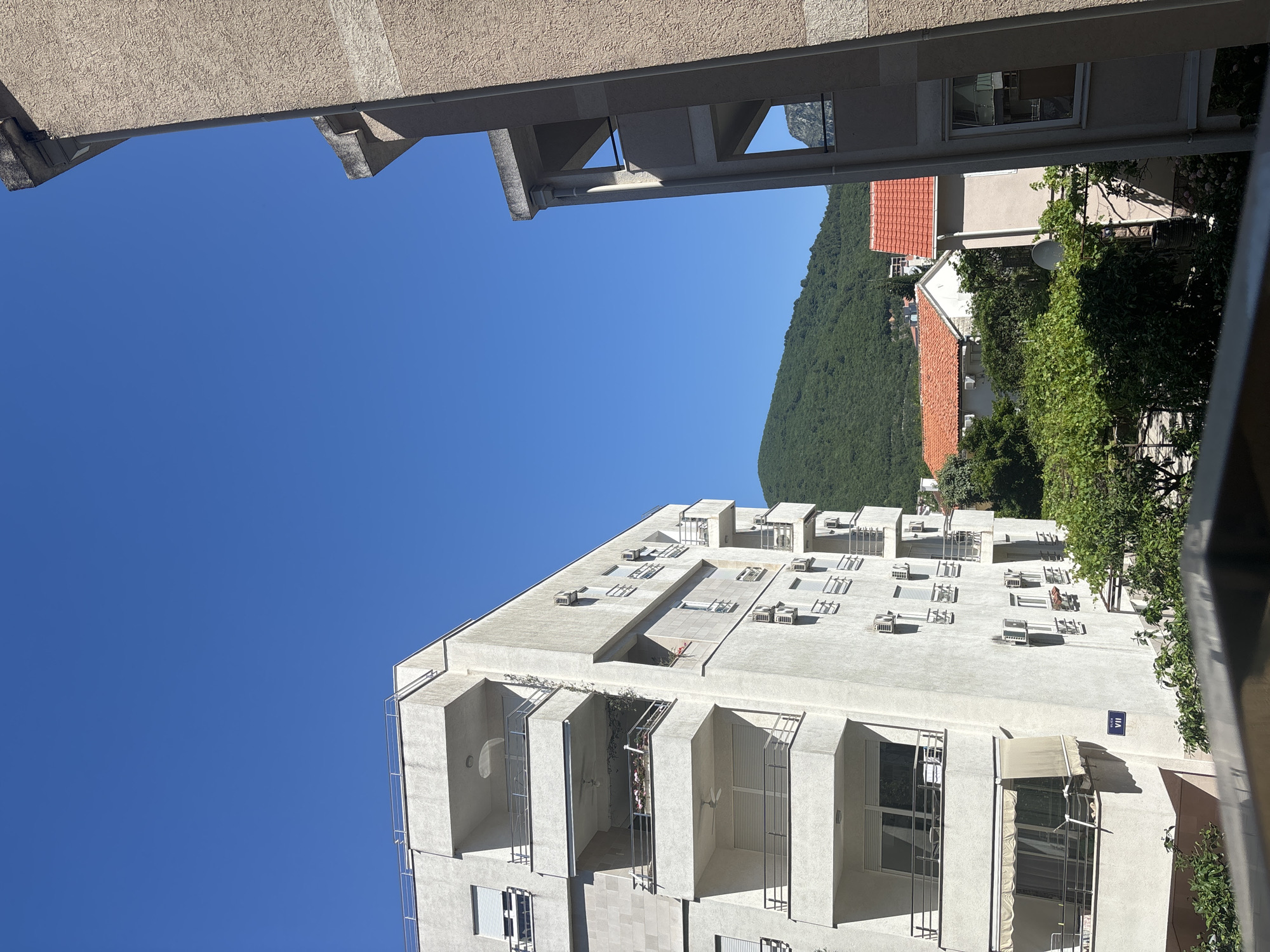 The image shows a view from between two buildings in Budva, Montenegro, with a clear blue sky dominating the upper left portion. To the right, behind the buildings, a green, tree-covered mountain rises. Lower in the image is a modern, white apartment building with a unique design featuring protruding balconies and numerous windows, each with a set of air conditioning units above. The rooftops are flat, with visible mechanical equipment. In the upper right, portions of other buildings are visible, including a red-tiled roof and some greenery. The overall impression is a bright, sunny day in a densely built urban environment surrounded by natural features.