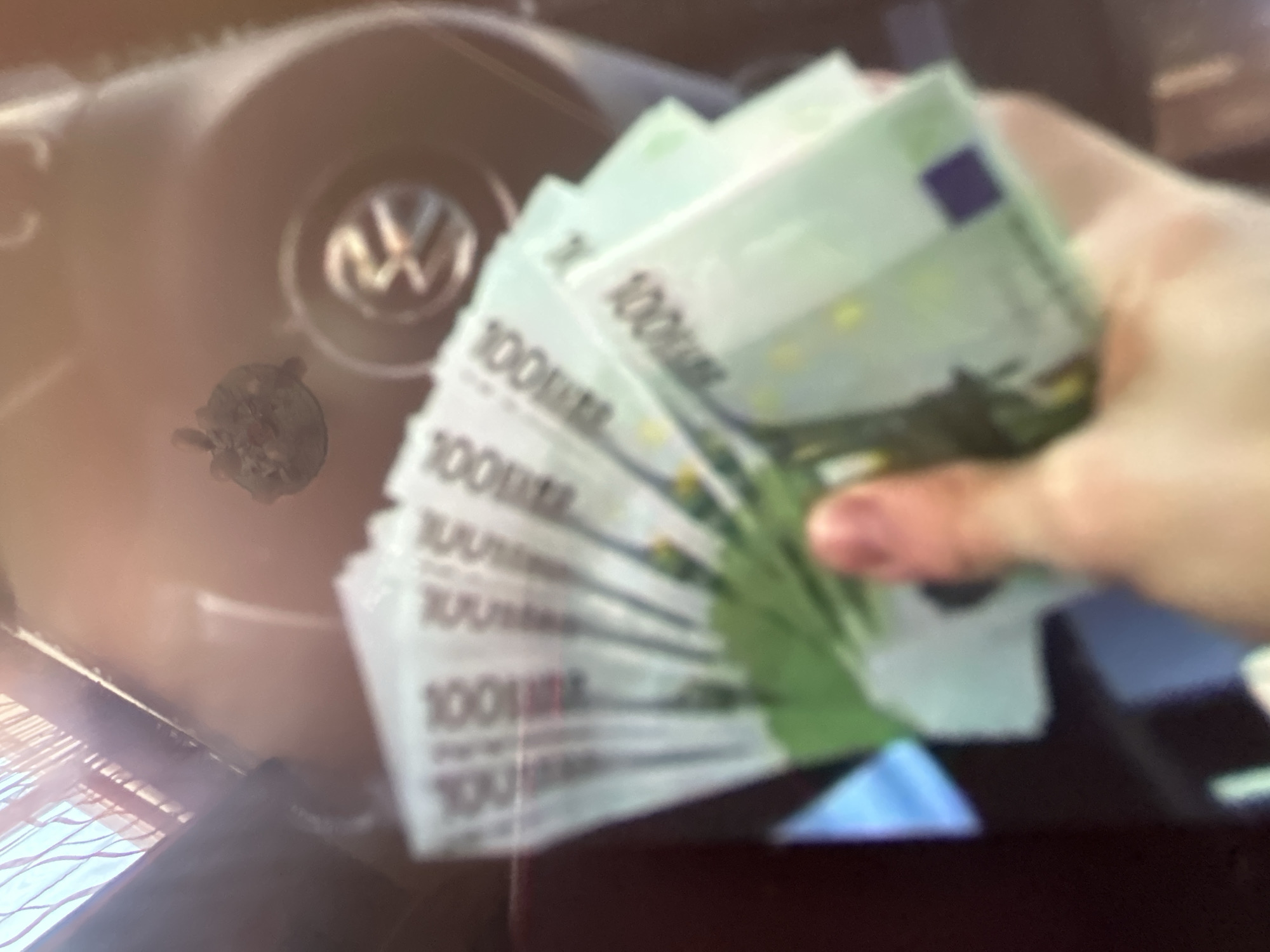The image shows a close-up of a hand holding several 100 Euro banknotes, fanned out in a semi-circle. The banknotes are mostly green and white with visible numbering and what appears to be a blue square on one of them. The hand holding the money is blurred, suggesting a focus on the currency itself.
In the background, out of focus, is a car's steering wheel with a Volkswagen logo in the center. To the left of the steering wheel, there is a small, dark, circular object with some detailing, possibly an air freshener or a small keychain. The overall scene appears to be inside a car, indicated by the steering wheel and the interior lighting. The lighting in the image is warm and diffused, with some bright reflections, suggesting it could be daytime. There are no clear indicators of the weather or specific time of day beyond that. The presence of money, combined with a car interior, could imply travel, a transaction, or simply displaying wealth. The image lacks any discernible emotional cues due to the focus on objects and the blurred nature of the human element. The visible text on the banknotes clearly reads "100 EURO". There are no other significant textual elements visible in the frame. The location context provided is Gelendzhik, Russia, however, there are no visual cues within the image to confirm this specific location.
