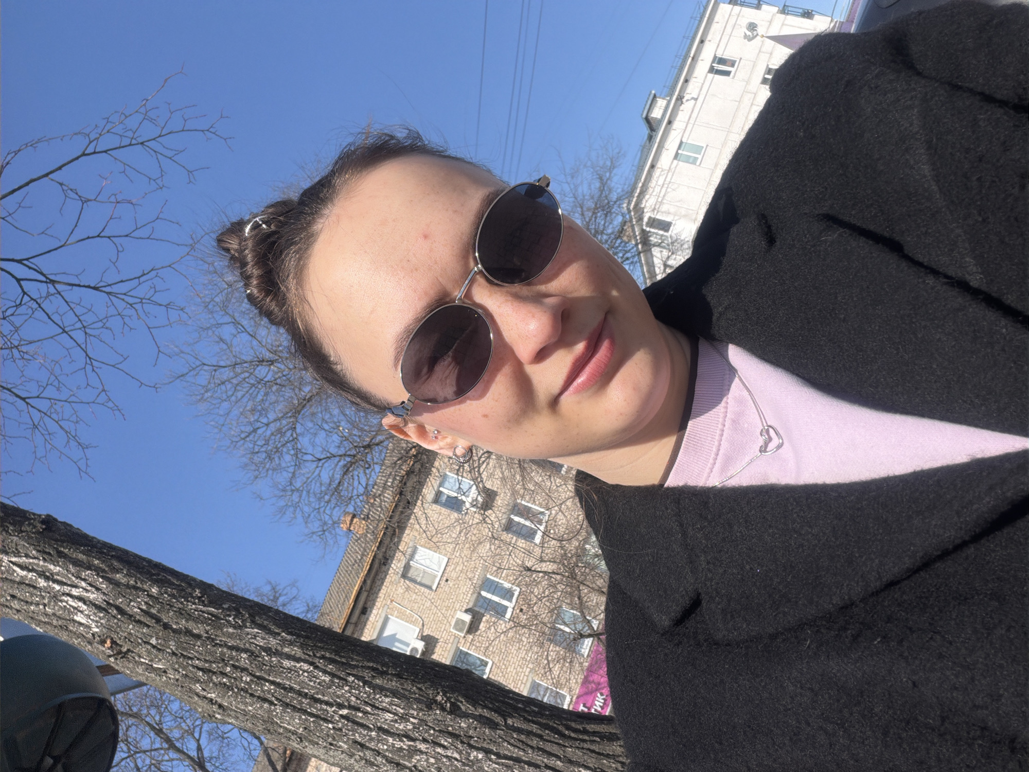 An outdoor, eye-level shot captures a female individual in the foreground within the city of Chaikovsky, Russia, under a clear blue sky. The individual, positioned slightly off-center and angled, wears dark oval sunglasses with thin silver frames, a light pink crew-neck top, and a black coat. A thin silver chain necklace with a heart-shaped pendant is visible. Her dark hair is pulled back into a bun secured with silver-colored clips. Multiple piercings are present on her left ear. She exhibits a subtle smile, looking directly towards the camera. To the left, a large tree trunk with dark, textured bark stands vertically, with bare branches extending into the upper frame. In the background, two multi-story buildings are visible. The left building features light brown brickwork, multiple windows, and a partial pink sign with Cyrillic text ("ТИК"). The right building is light-colored, possibly white or light grey, also featuring multiple windows. Bare tree branches are scattered across the mid-ground. A dark blue object, possibly part of a street light, is partially visible at the top-left behind the tree. The scene is illuminated by bright sunlight, indicative of daytime.