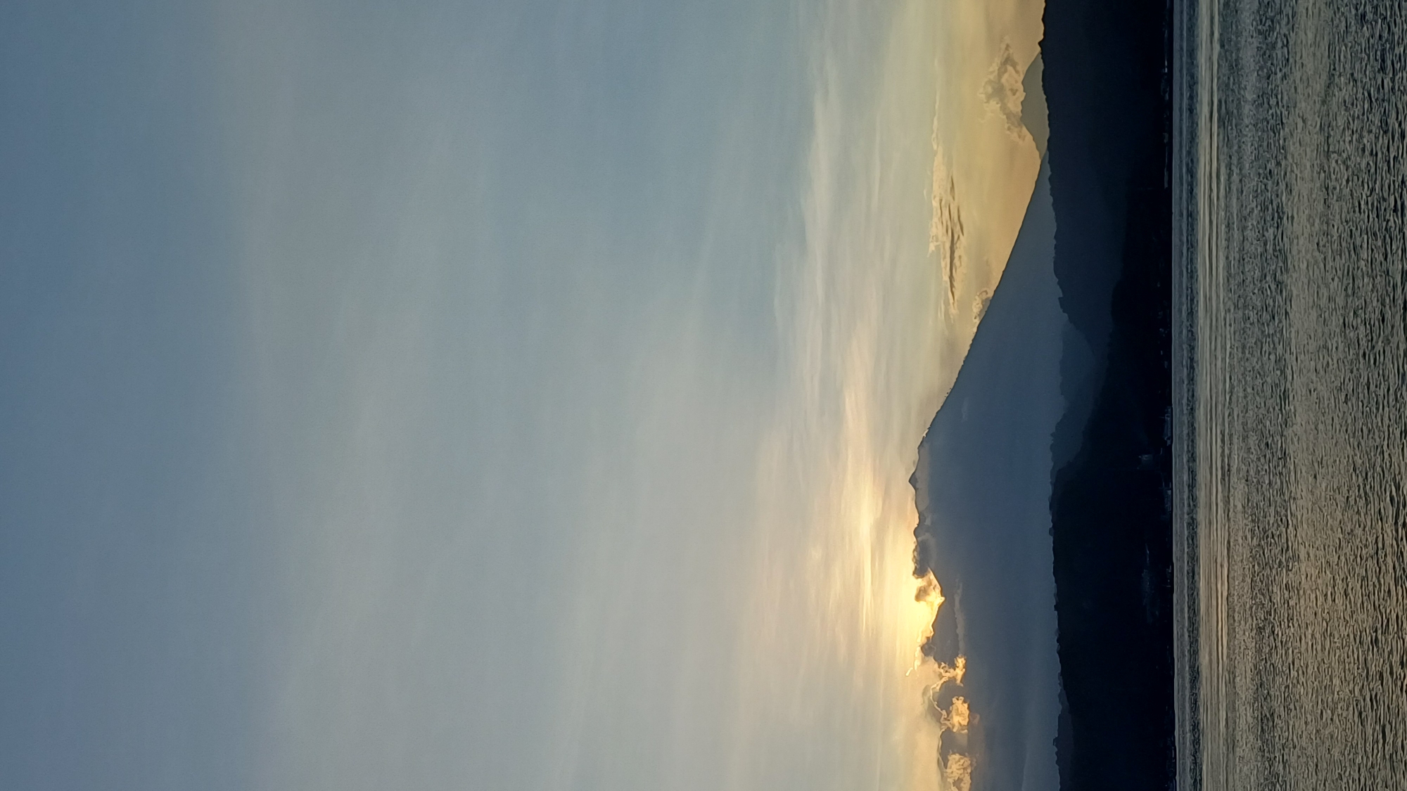 The image shows a landscape with a mountain range silhouetted against a sky transitioning from light blue to a yellowish-golden hue, likely at sunrise or sunset. A body of water reflects the light, showing ripples and variations in tone. The location is in the city of Unknown, Indonesia.