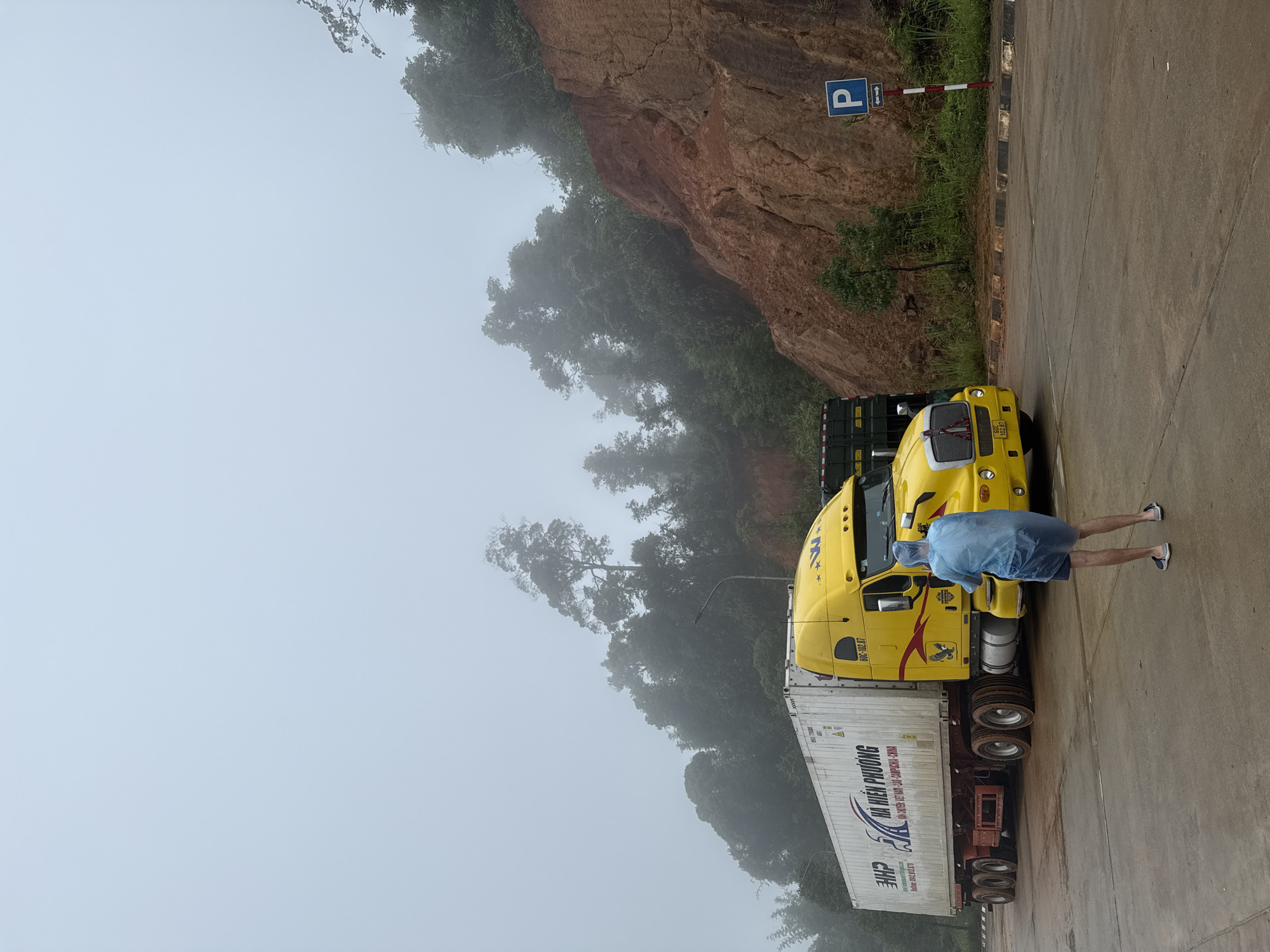 The image shows a bright yellow semi-truck with a white shipping container parked on a wet, paved surface, likely a road or parking area. The truck has Vietnamese license plates that read "60C-102.87". A person wearing a light blue poncho and shorts stands facing the truck, holding what appears to be a phone or camera. The environment is characterized by a steep, reddish-brown cliff face behind the truck, covered in green vegetation. The sky is overcast and foggy, creating a hazy atmosphere and obscuring the upper portions of the trees. A blue parking sign with an arrow pointing left is visible on the right side of the image. The overall impression is a roadside scene in a rural or mountainous area. The presence of Vietnamese license plates and the distinct landscape suggest the location is likely within Vietnam, possibly near Bo Y, which is known for its mountainous terrain and border crossings.